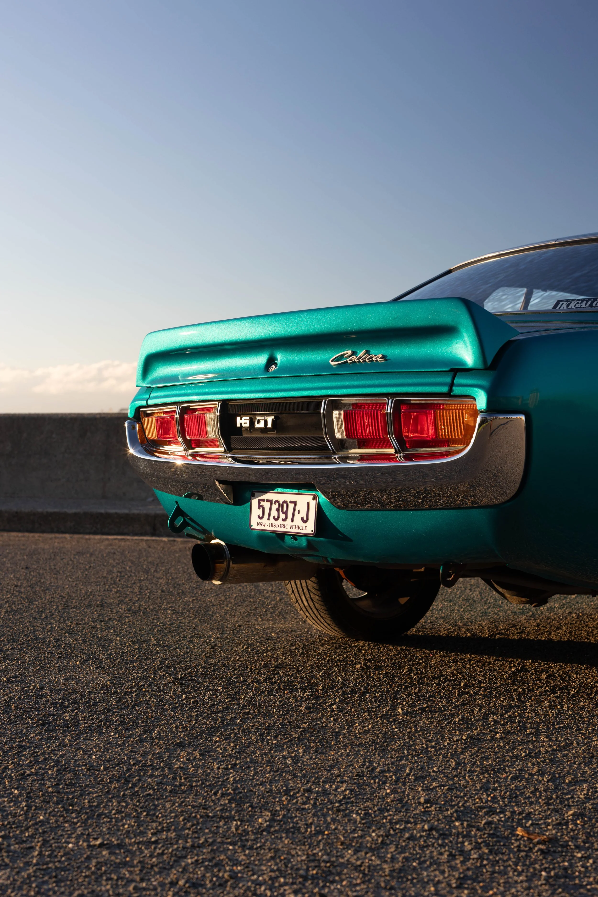 Toyota Celica 1600GT rear three-quarter at golden hour, ducktail boot lip, GT tail-light panel and stainless exhaust tip classic car detail photography.