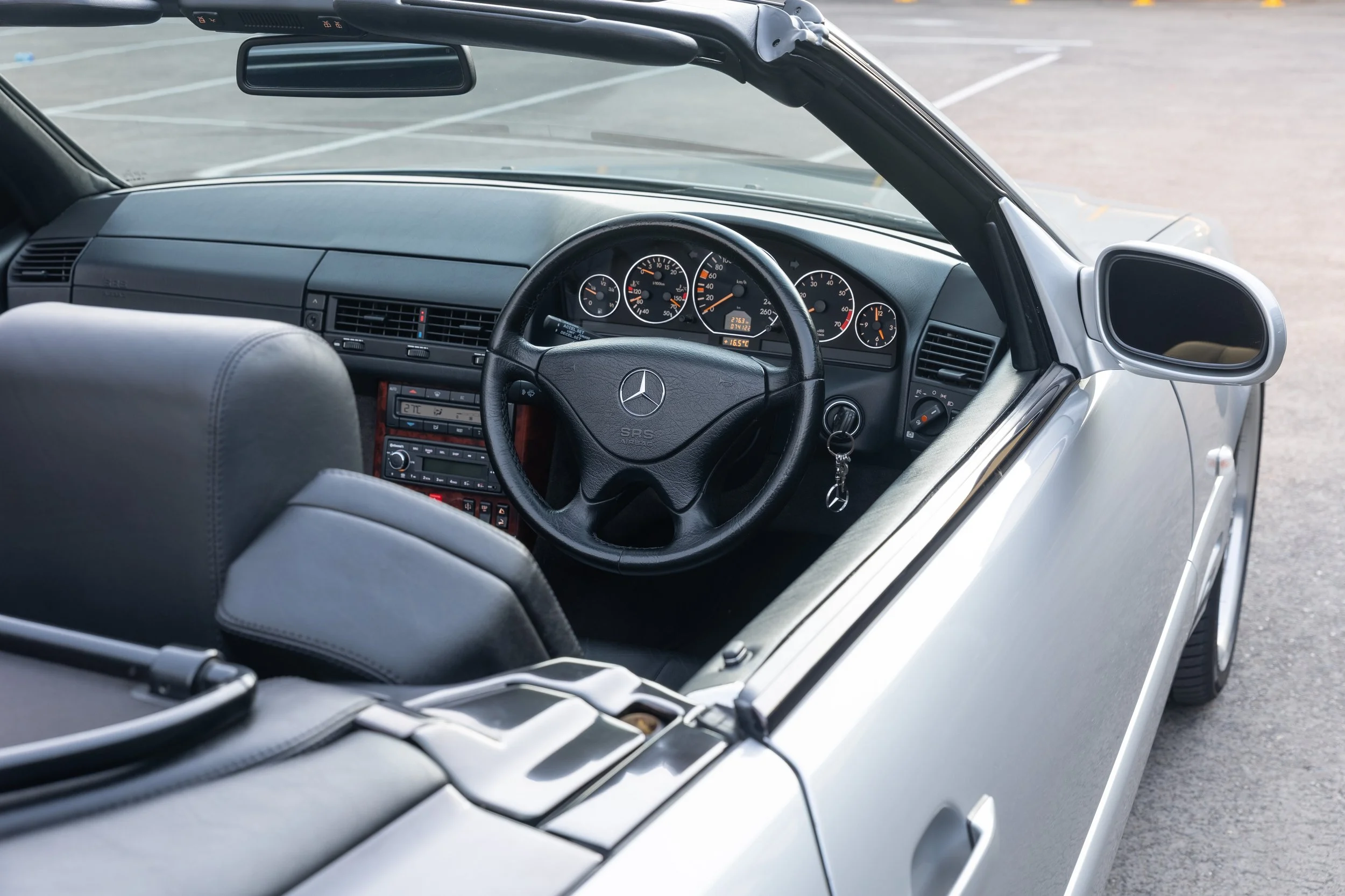 Interior of a vintage convertible R129 Mercedes-Benz car, showing the dashboard, steering wheel, and rearview mirror, parked in an empty parking lot.