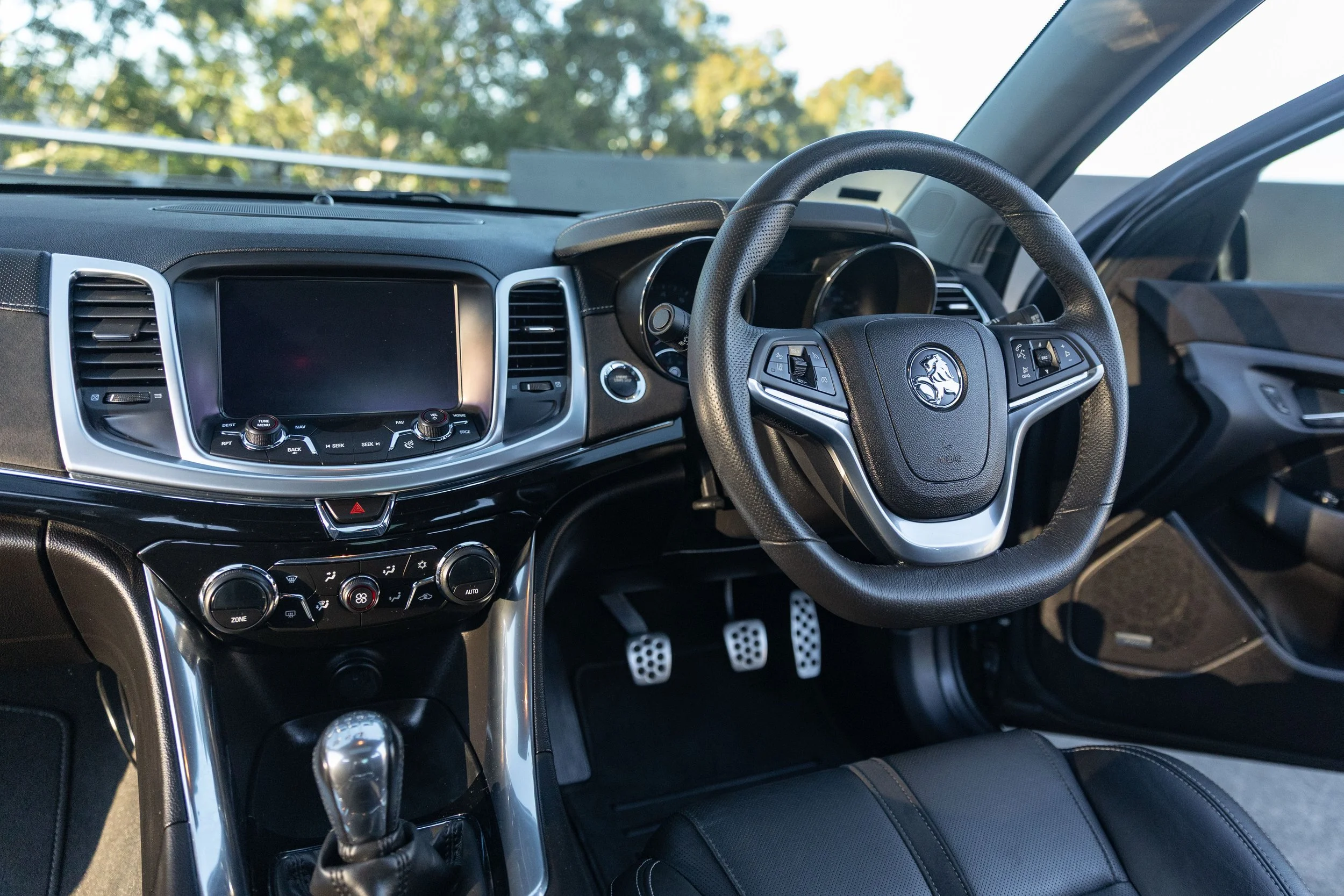 Interior view of a car dashboard and steering wheel, with a view of the windshield showing trees outside. The steering wheel has a logo of a lion, and the dashboard features various control buttons and a display screen.