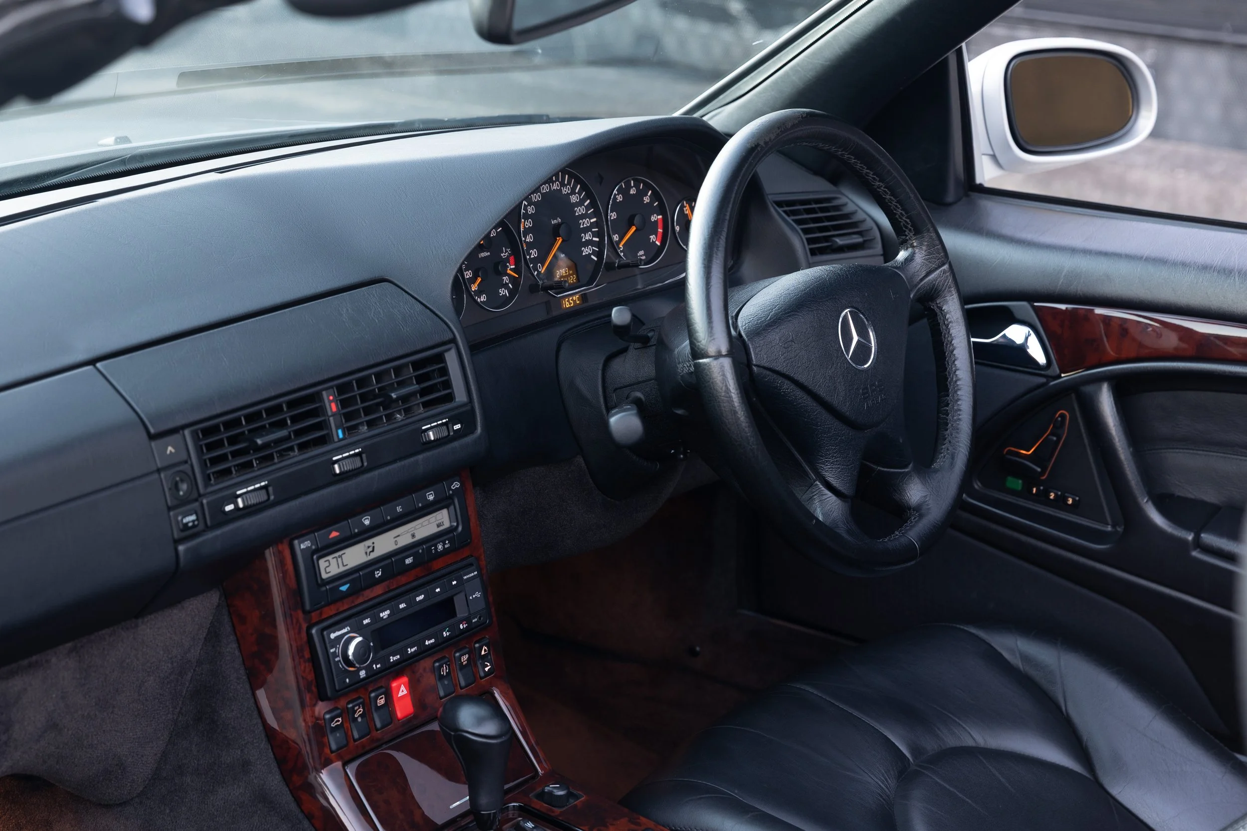The interior of a silver R129 Mercedes car showing the dashboard, steering wheel, and gear shift. The dashboard features a speedometer, tachometer, and other gauges, with a climate control panel below. The steering wheel has the Mercedes-Benz logo.