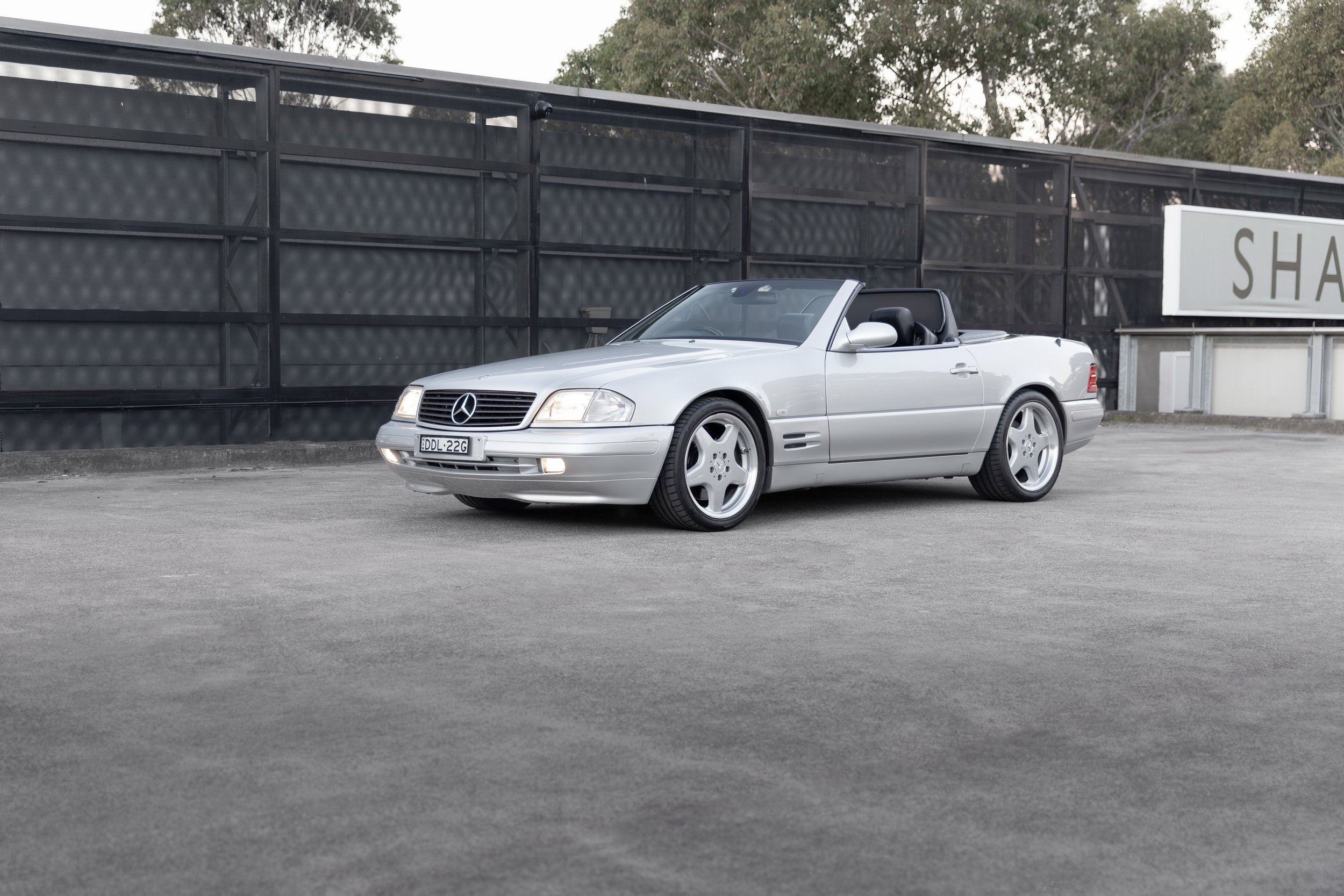 A silver R129 SL 500 convertible Mercedes-Benz parked in an outdoor lot with a black fence and trees in the background.