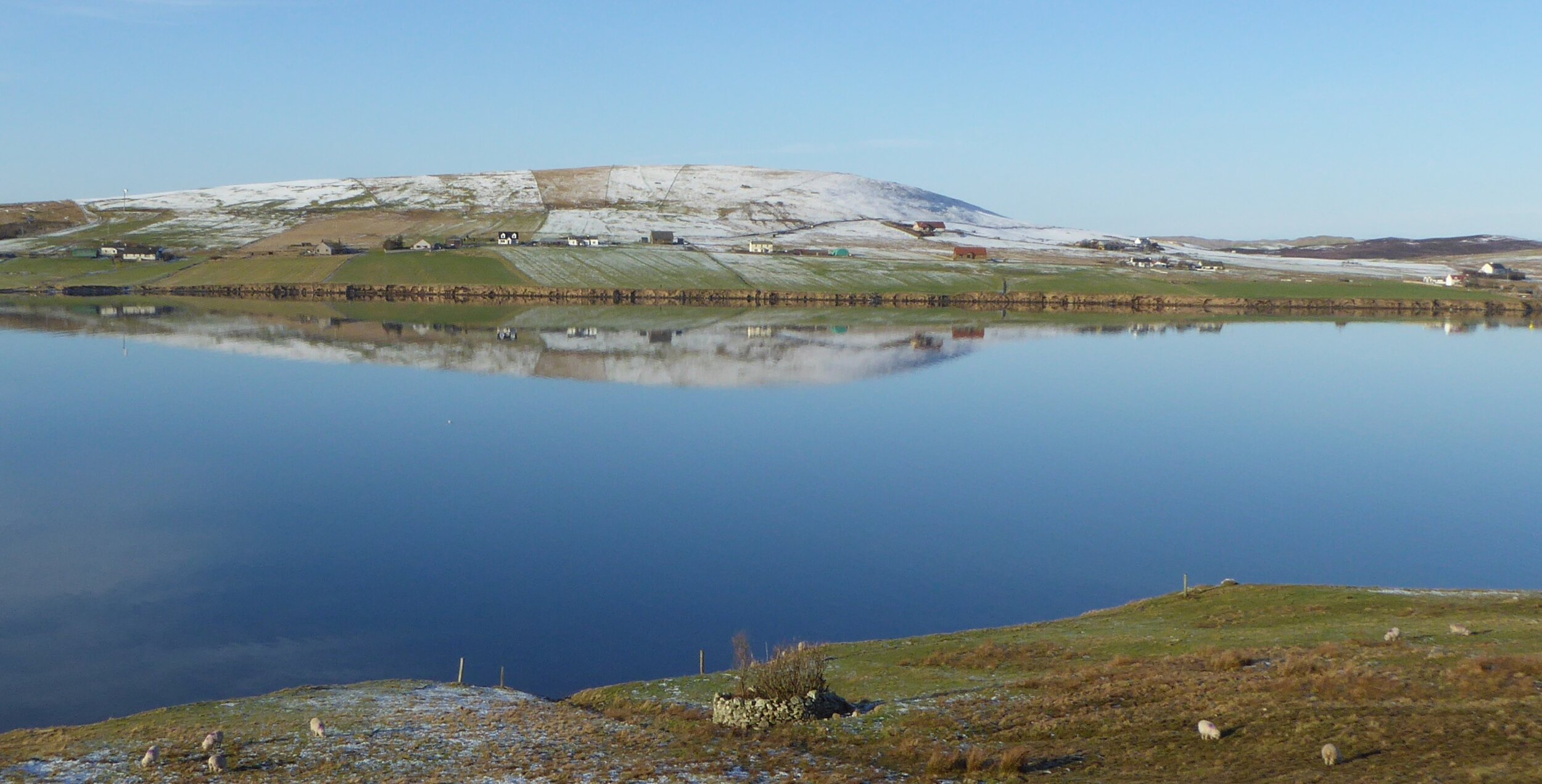 A still day in Aith where the low sun and reflections really bring out the way communities are structured: the houses run along the edge of the water, with the parks, the cultivated fields, running down to the water, and the scattald, or shared hill 