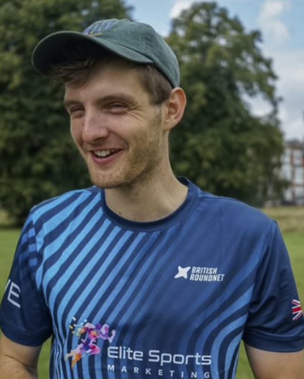 Young man smiling outdoors, wearing a blue sports jersey and a gray cap, with trees in the background.