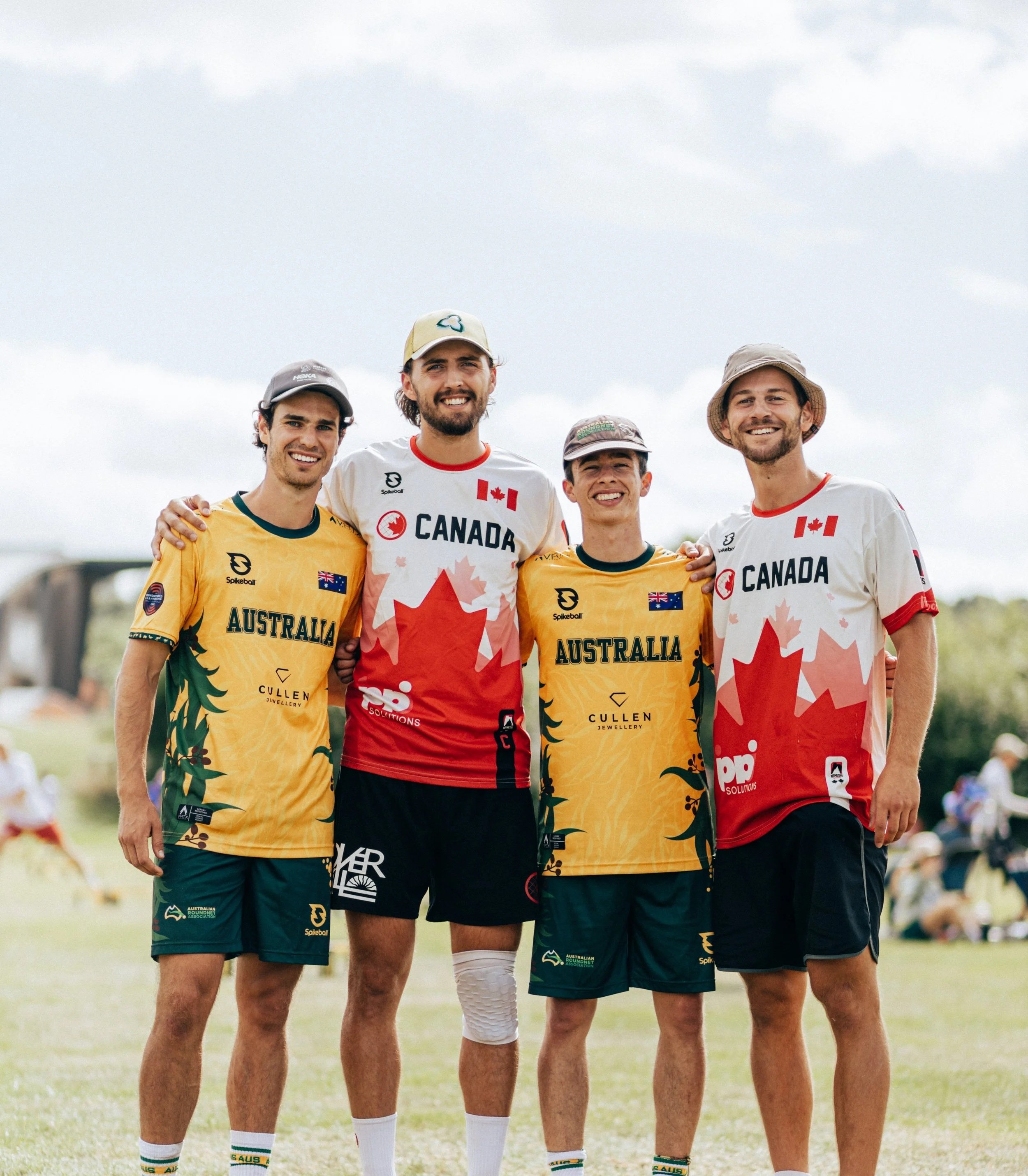 Four male athletes standing outdoors on grass, smiling, with arms around each other. They wear team jerseys for Australia and Canada, indicating a sports event.