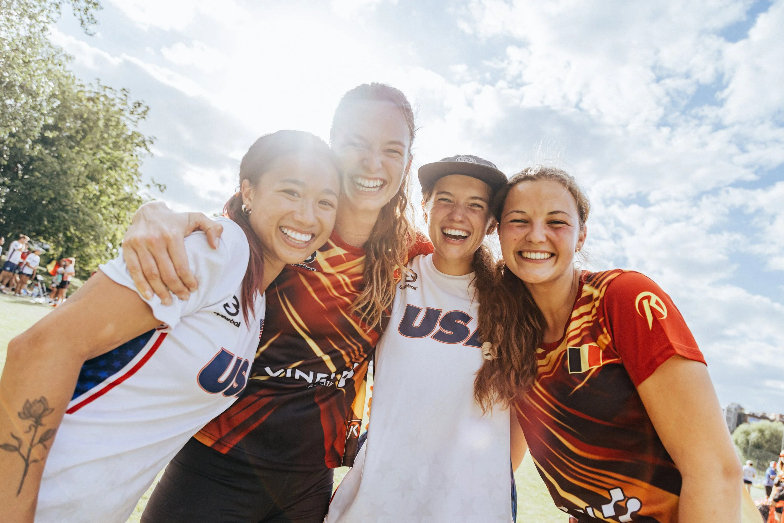 Four female athletes smiling and huddling outdoors during daytime, wearing sports jerseys with 'USA' on them, with a crowd and trees in the background.