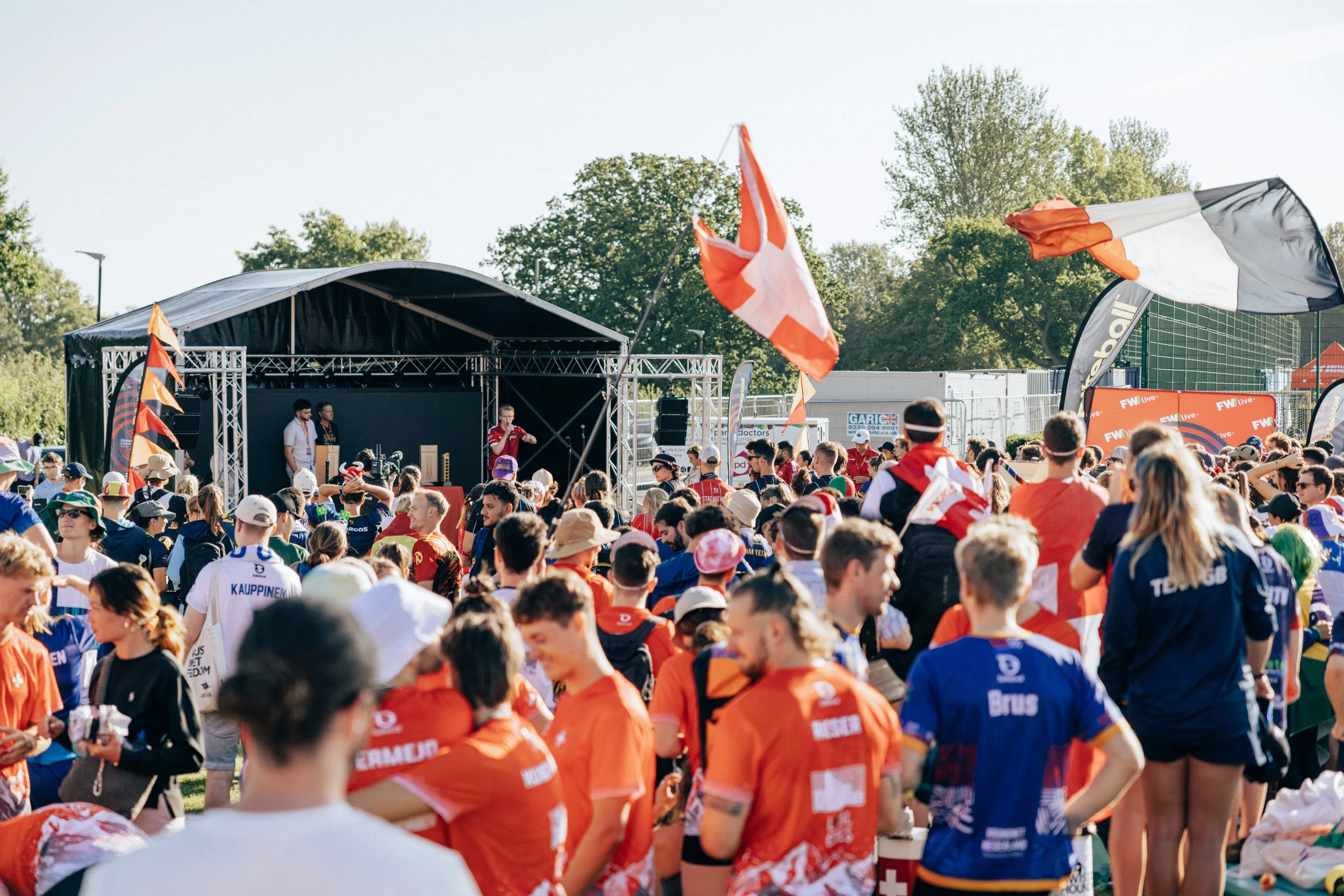 Large outdoor gathering of sports fans, many wearing jerseys and hats, attending an event with flags waving, a stage with speakers and performers, and trees in the background on a sunny day.