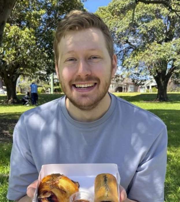 A smiling young man with light brown hair and a beard holding a box of baked goods outdoors in a park with trees and a person in the background.