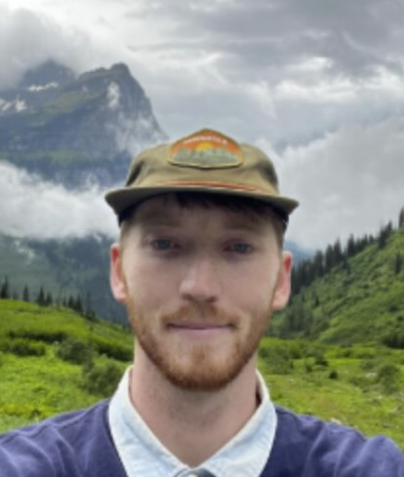 Young man with red hair and beard wearing a baseball cap and white shirt in a lush, green mountainous landscape with cloudy sky.