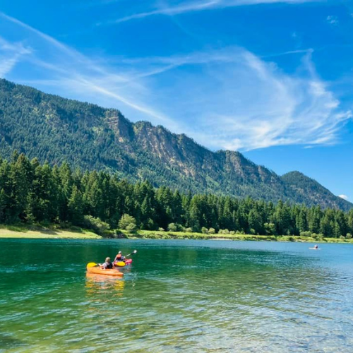 two people in kayaks on a turquoise colored river with a mountain ridge in the background and blue sky