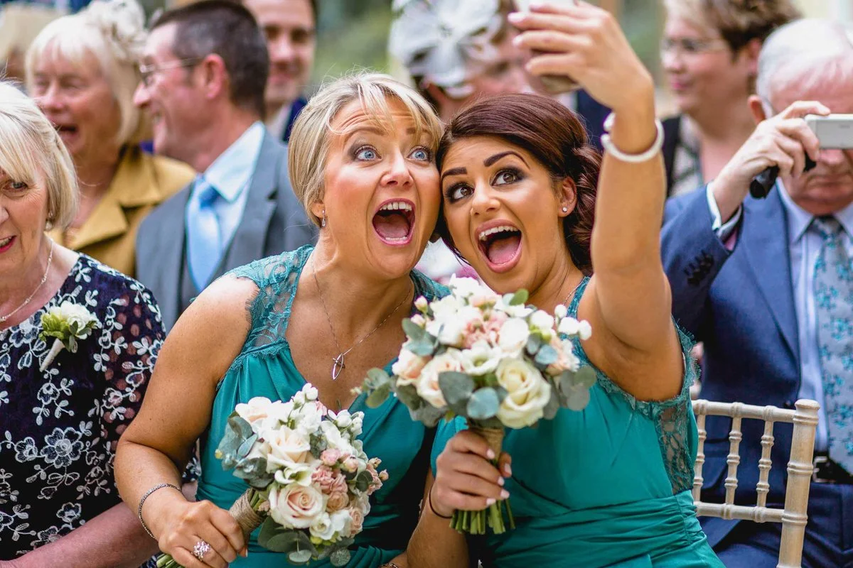 two bridesmaids pose for a selfie during a wedding ceremony, they wear jade dresses and each hold bouquets of roses and eucalyptus whilst their attention is towards the phone which one of them is holding up