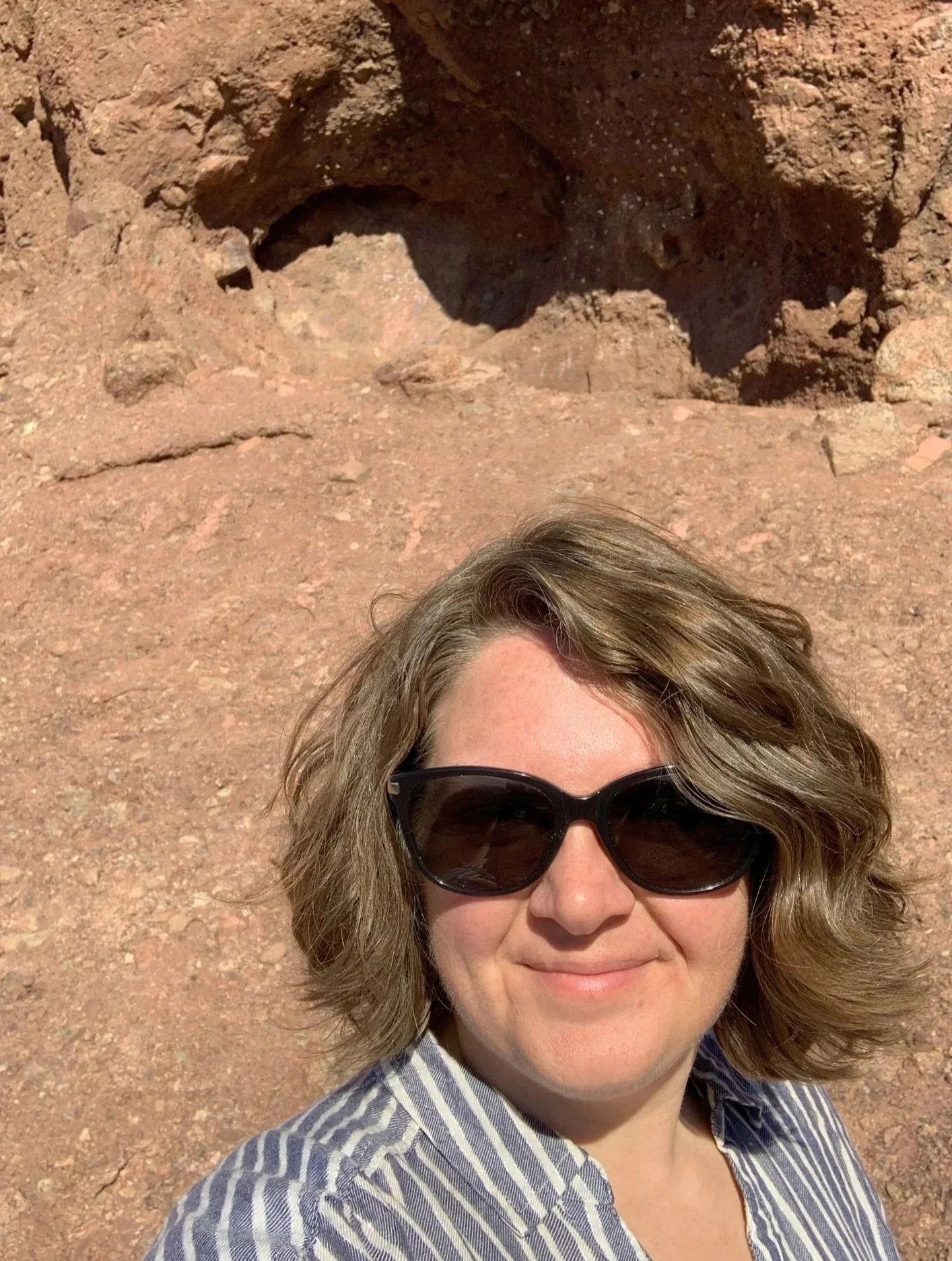 A woman with wavy blonde hair wearing large sunglasses and a blue and white striped shirt, smiling in a desert environment with reddish-brown soil and rocks.