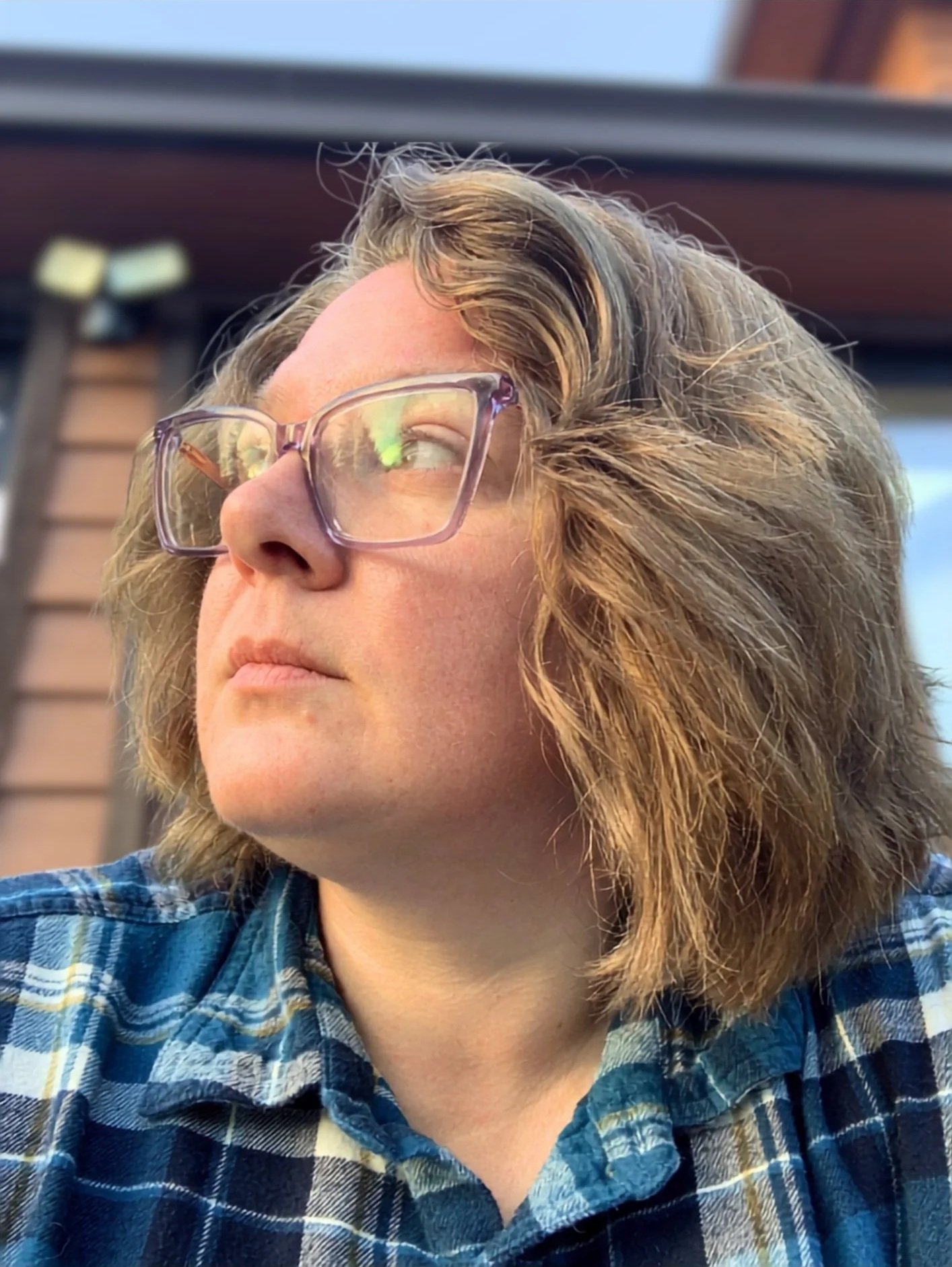 Close-up of a woman with blonde hair wearing glasses and a blue plaid shirt, looking into the distance outdoors.