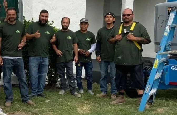 Group of nine men standing outdoors in front of a white building with a large rounded window, wearing matching dark green T-shirts, some giving thumbs-up, smiling.