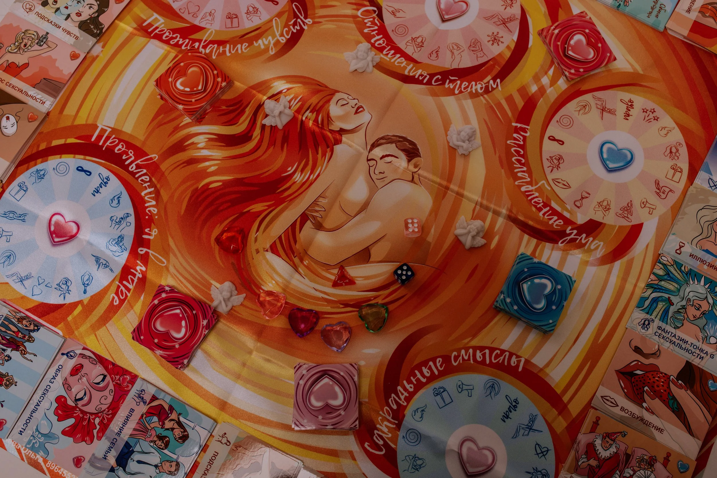 A colorful board game with illustrations of romantic and sensual themes, featuring hearts, flowers, and images of a woman with long red hair. There are game pieces shaped like hearts, some dice, and various cards on the game board.