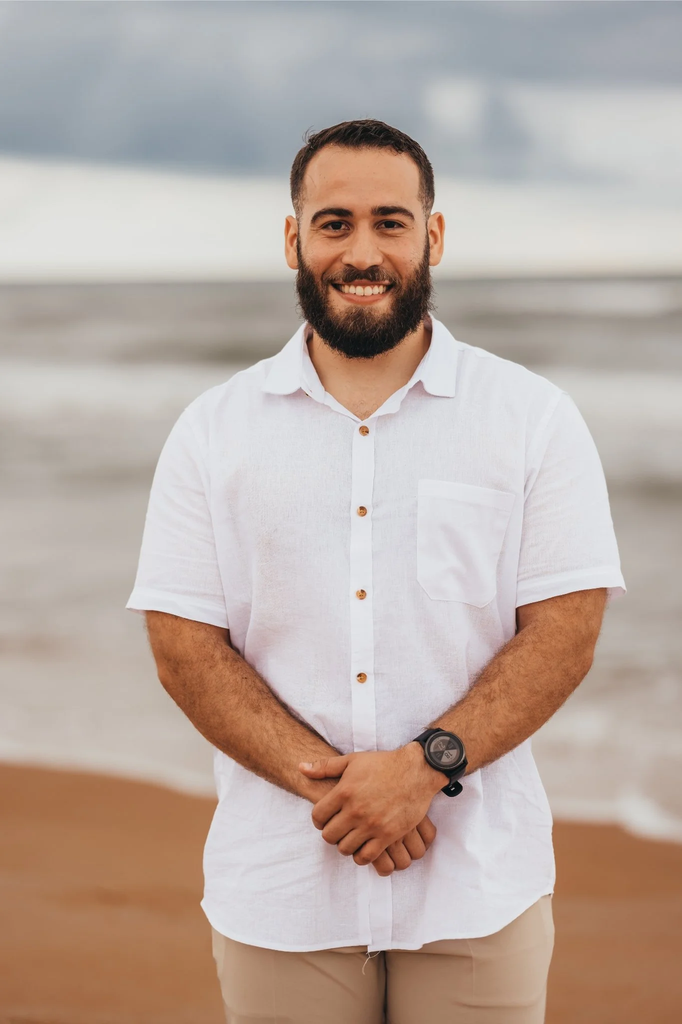 A smiling man with a beard and short hair standing on the beach during overcast weather, wearing a white short-sleeve button-up shirt and a black wristwatch.