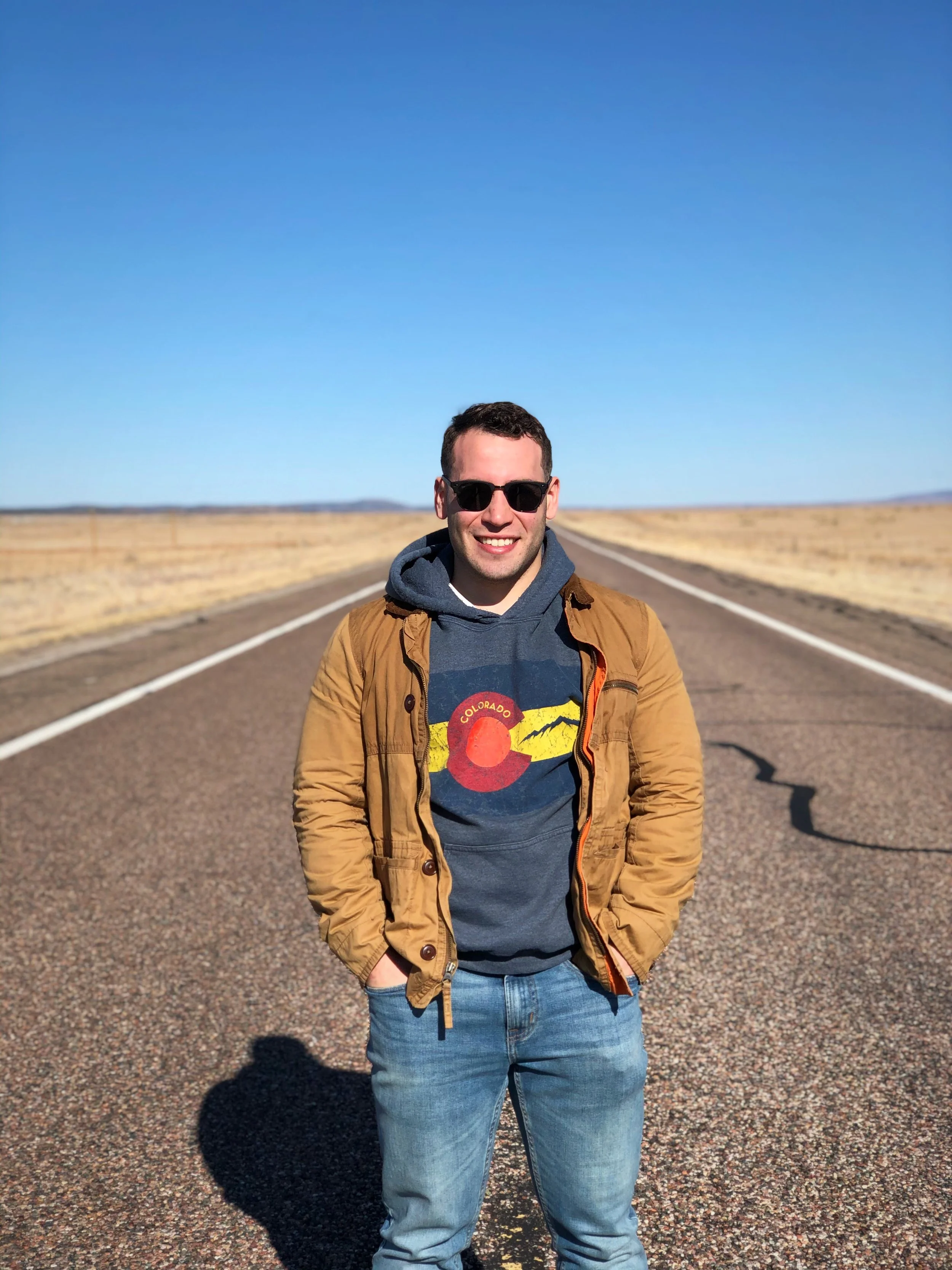 A smiling man wearing sunglasses, a Colorado-themed shirt, a tan jacket, and blue jeans standing on an empty desert road under a clear blue sky.