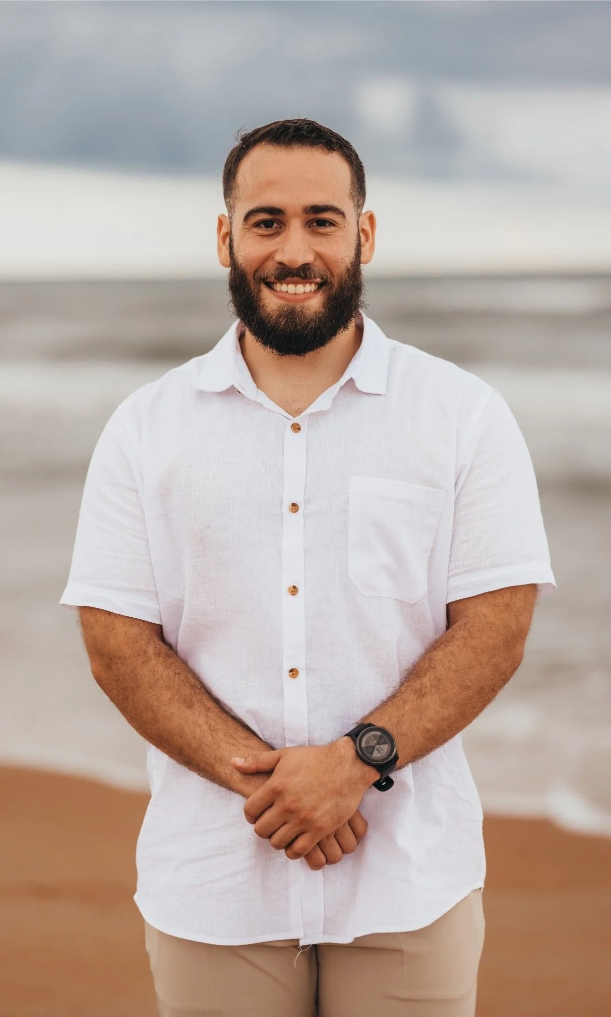 A man with a beard wearing a white shirt and beige pants, standing on a beach with the ocean and cloudy sky in the background.