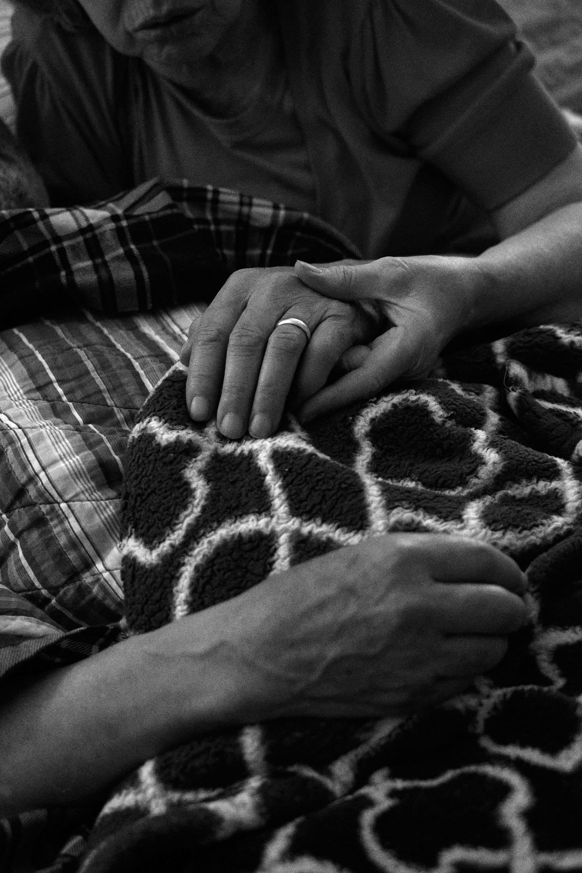 Black and white image of an elderly woman holding a man's hand, with a plaid blanket and patterned fabric in the background.