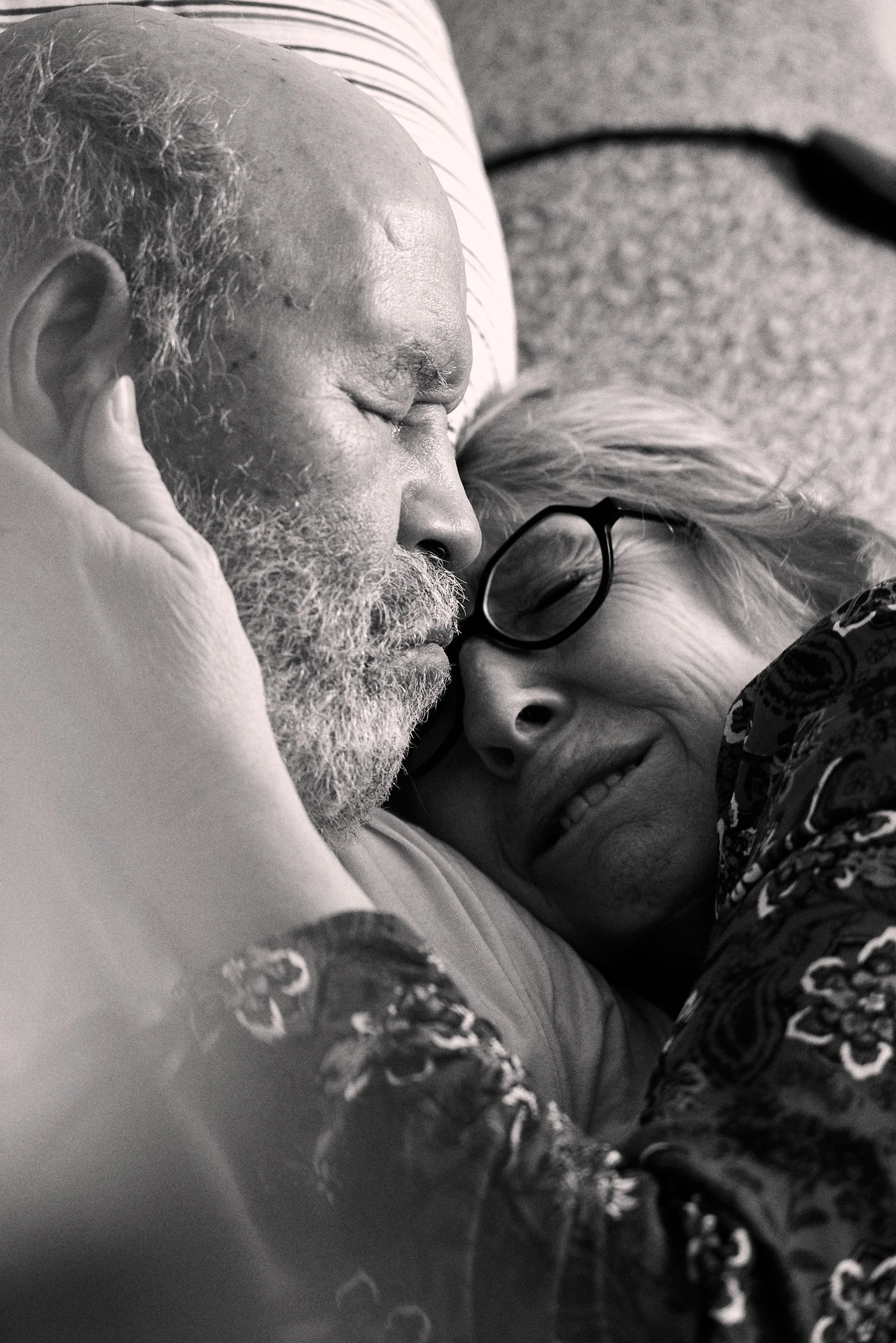 Elderly couple embracing closely, man with a beard and woman wearing glasses, displaying affection and contentment.