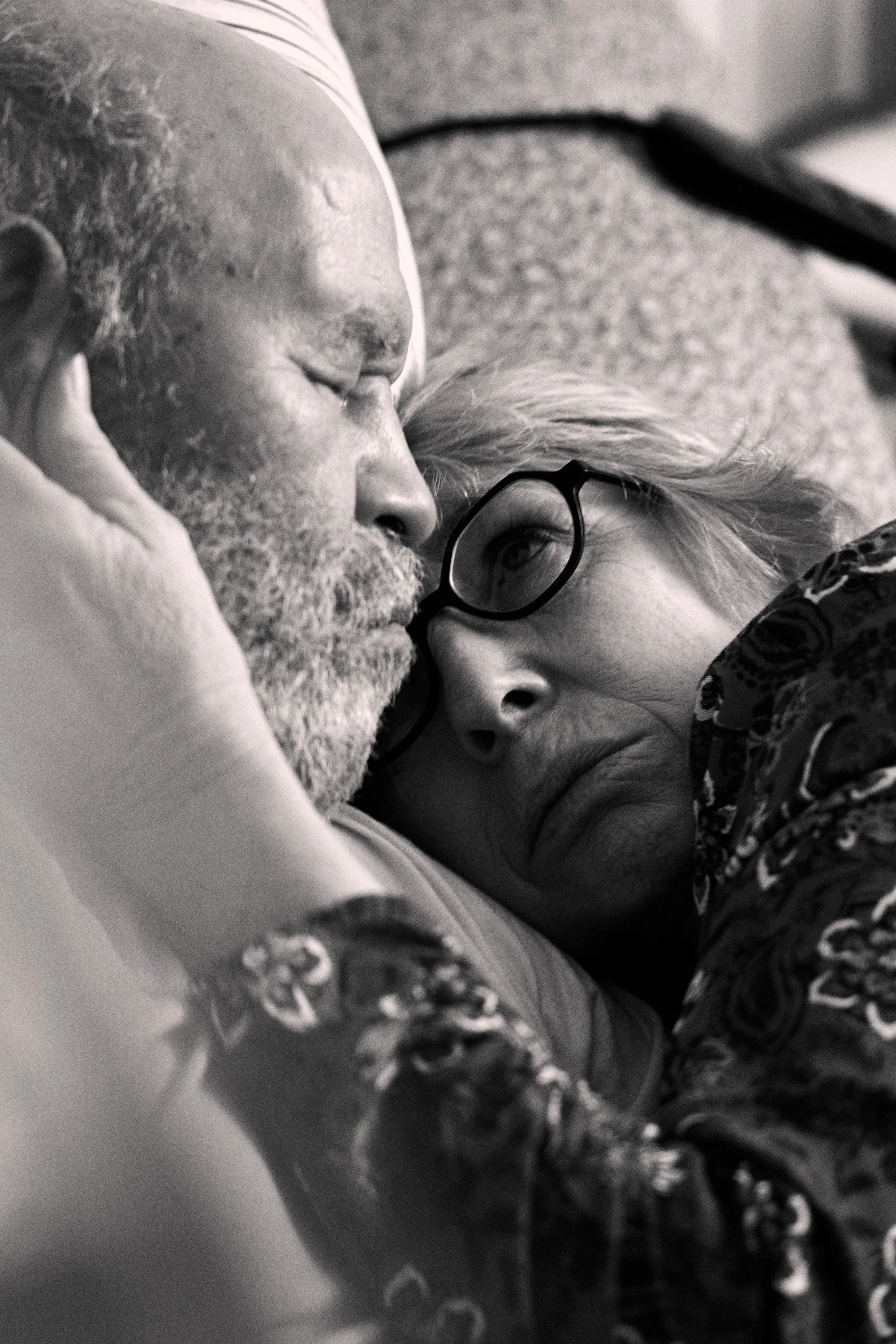 An elderly couple lying closely together, with the woman's head on the man's shoulder, both with a calm expression. The image is in black and white.