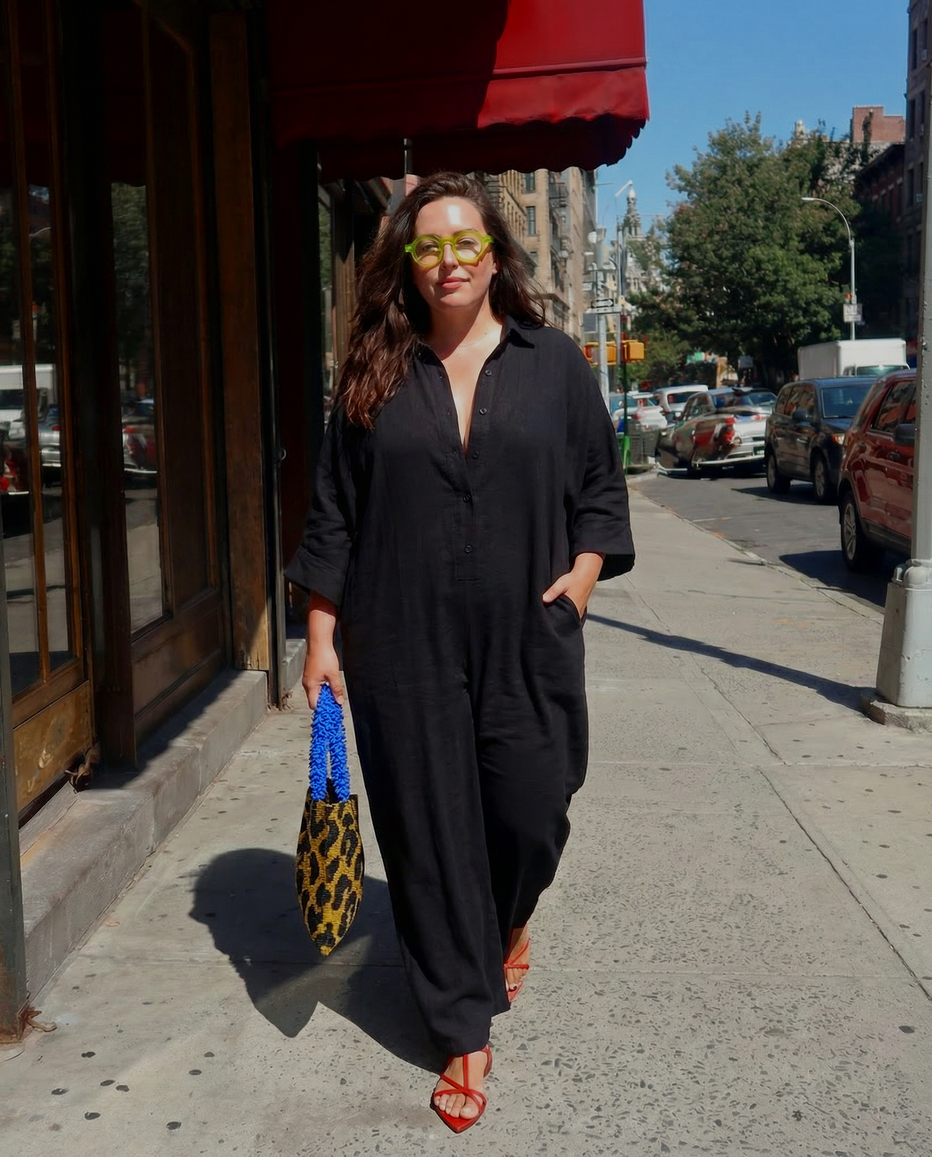 Woman walking on city sidewalk wearing black outfit, yellow glasses, red heels, carrying leopard print bag with blue handle, and sunglasses, under a red awning.