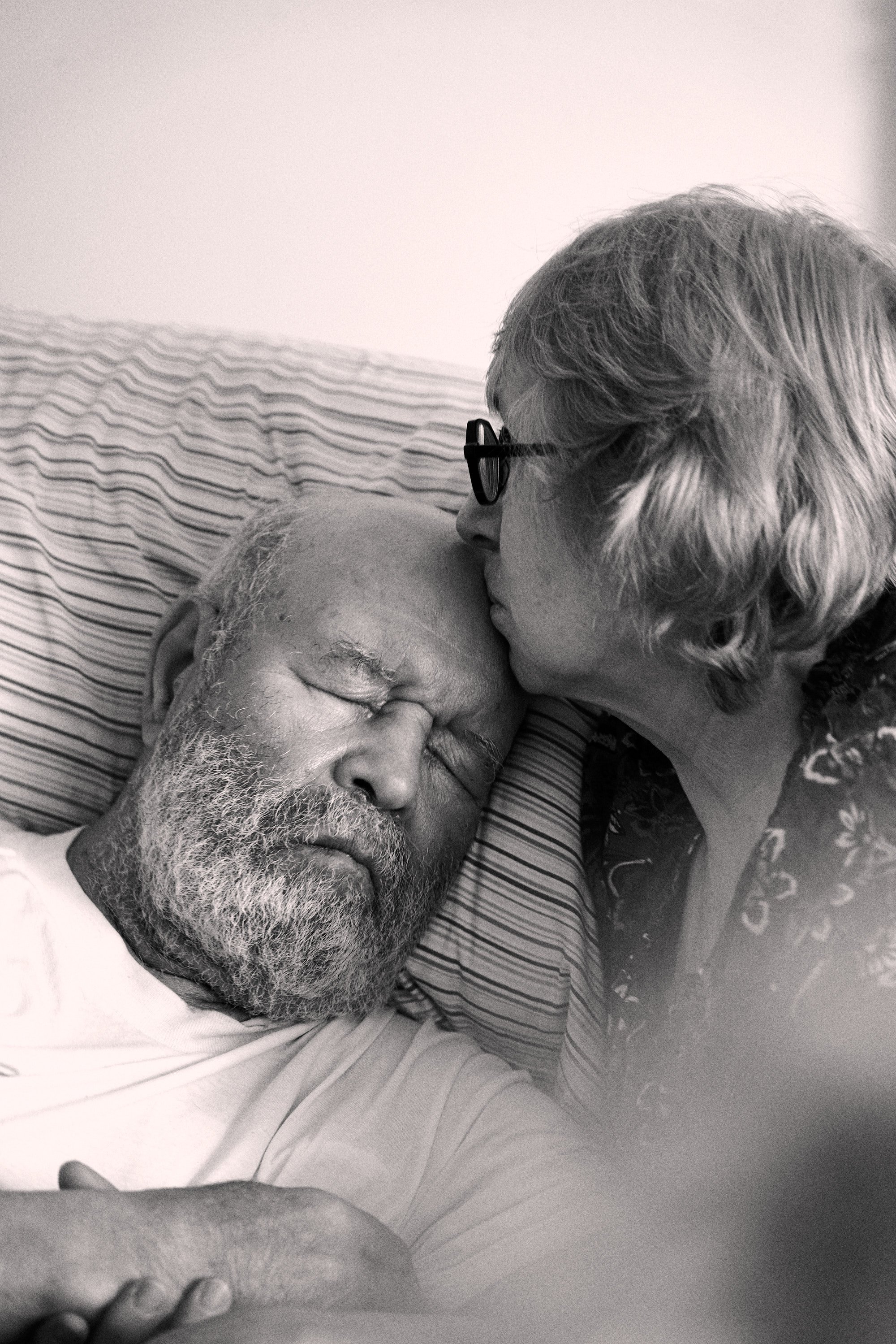 A black and white photo of an elderly woman kissing the forehead of an elderly man who is lying in bed with his eyes closed, depicted in a tender and intimate moment.