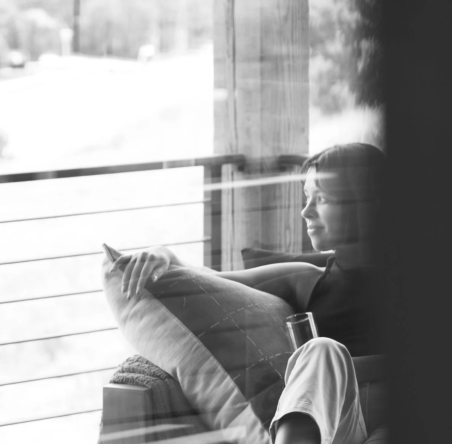 A woman sitting on a couch with a pillow, looking out the window with sunlight streaming in, holding a glass, in a cozy indoor setting.