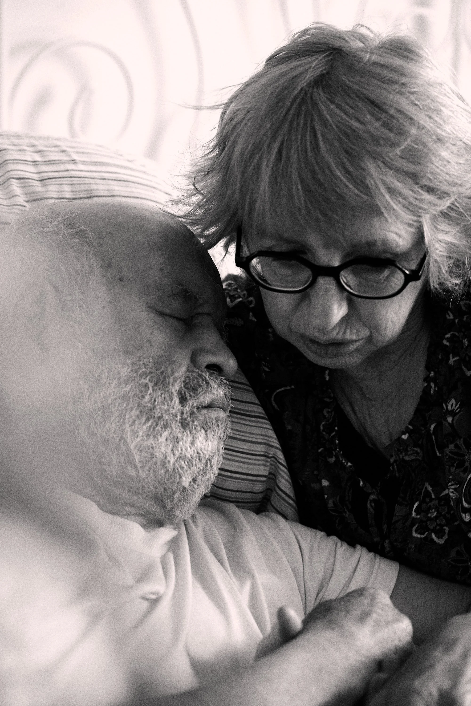An elderly man sleeping in bed with an elderly woman sitting beside him, looking down with concern. Black and white photograph.