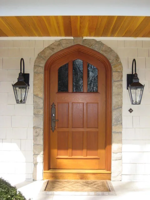 Wooden front door with three small windows at the top, framed by stone arch, flanked by two black outdoor lanterns, with beige brick wall and wooden ceiling above.