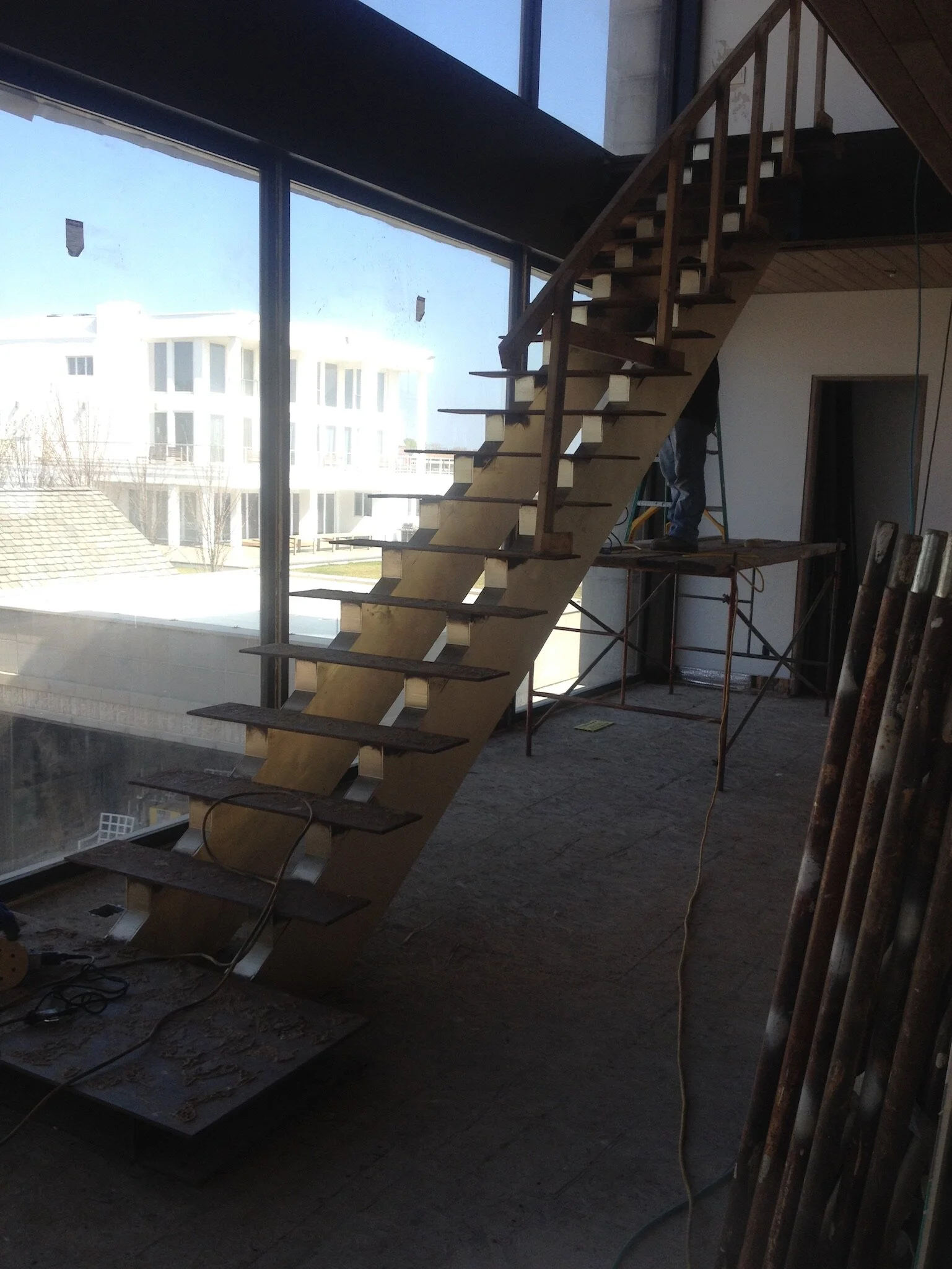 Interior of a building under construction, featuring a metal staircase without handrails, with large windows showing residential buildings outside, construction tools and scaffolding present.