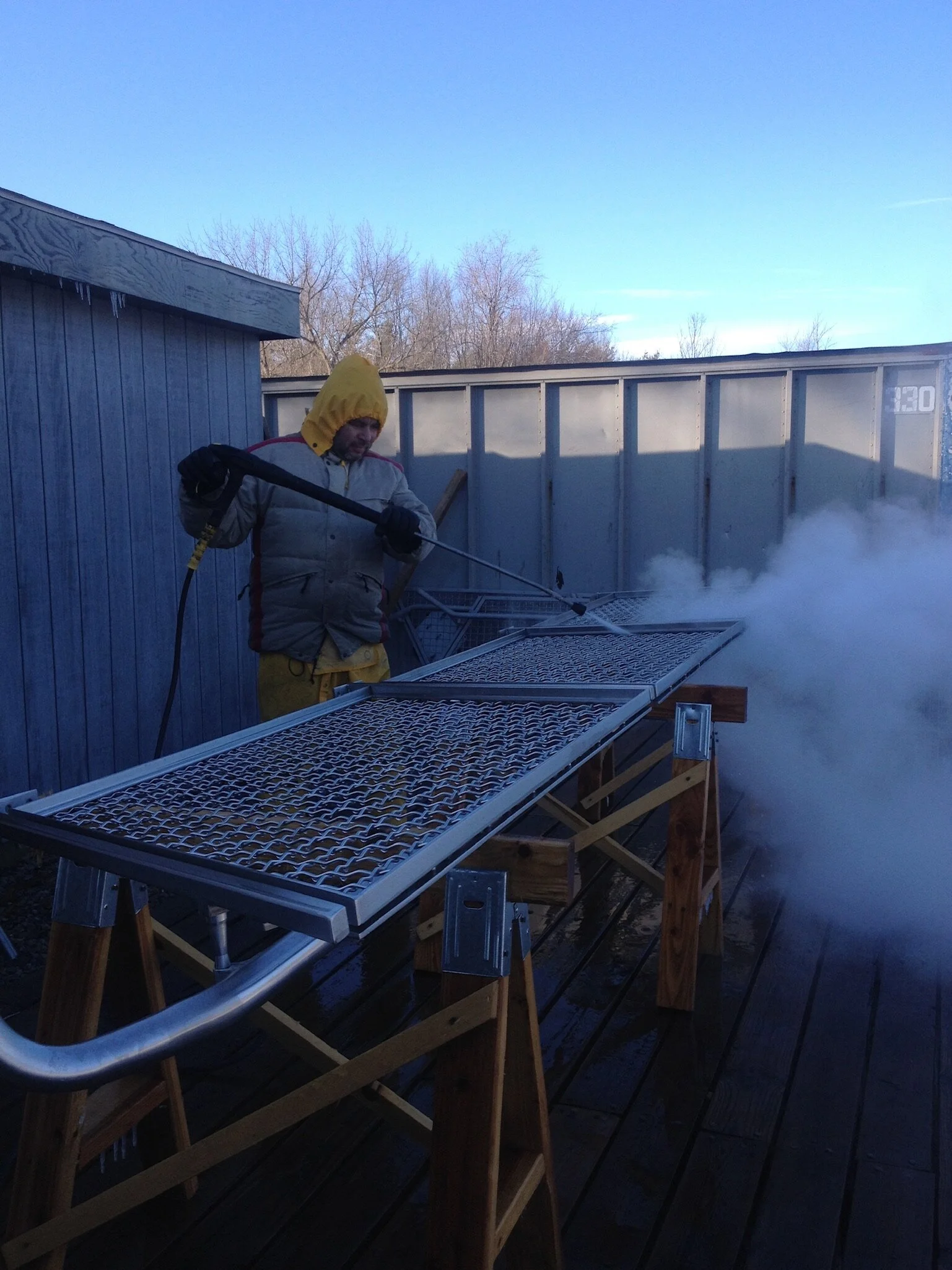 A person wearing winter clothing and a yellow hood is pressure washing a metal grate outdoors, creating a cloud of steam or spray. The scene is set on an outdoor deck with buildings and leafless trees in the background.