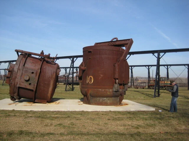 Two large, rusted industrial tanks or containers placed outdoors on a concrete pad, with a person standing nearby and a rail track in the background.