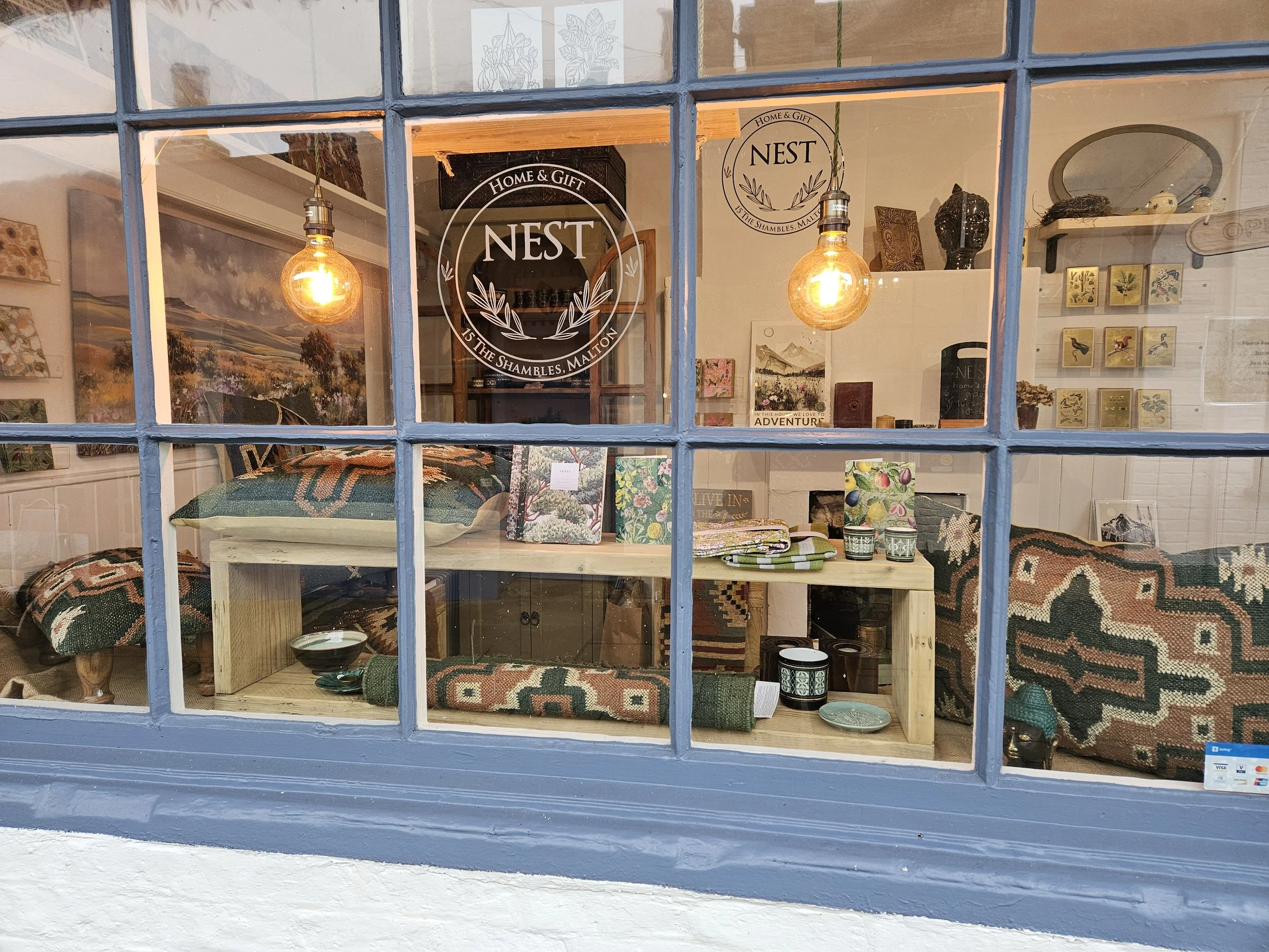 The front window of Nest Home and Gift shop in The Shambles. The windows are single glazed with blue frames looking into the softly lit gift shop with calm furnishings and gift cards.