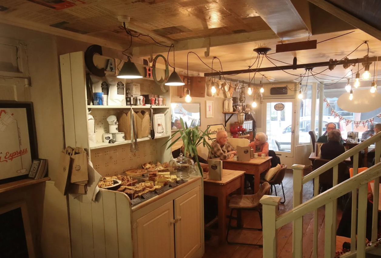 A warm and welcoming coffee shop with an older couple sat at a wooden table in the distance. Warm lighting and cakes on the dresser at the forefront.