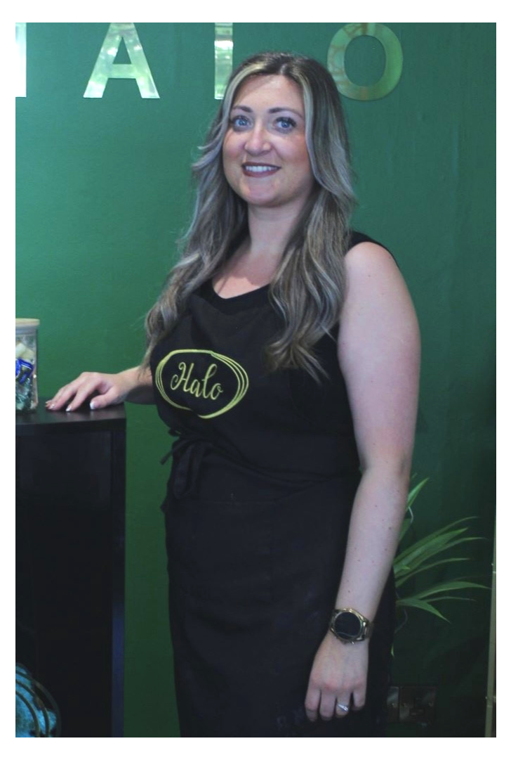 The owner is standing in the salon with a Halo branding top on infront of a green background.