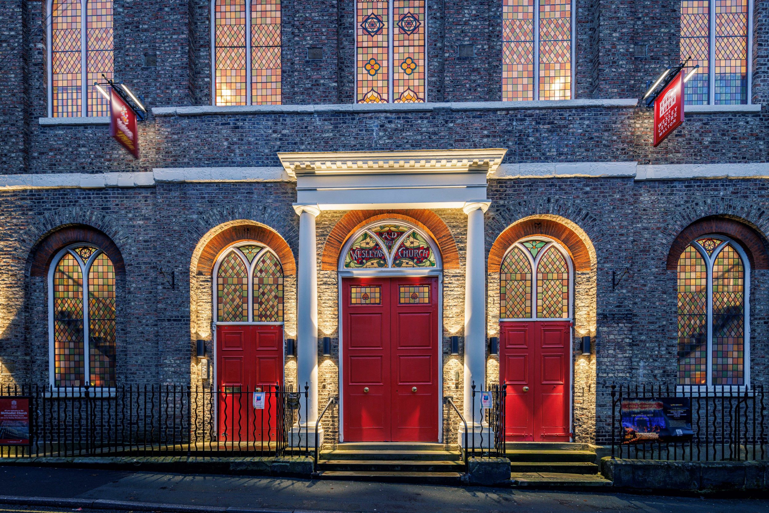 A front image showing the three arches of the red doors with intricate stained glass windows above at The Wesley Centre on Saville Street, Malton, North Yorkshire