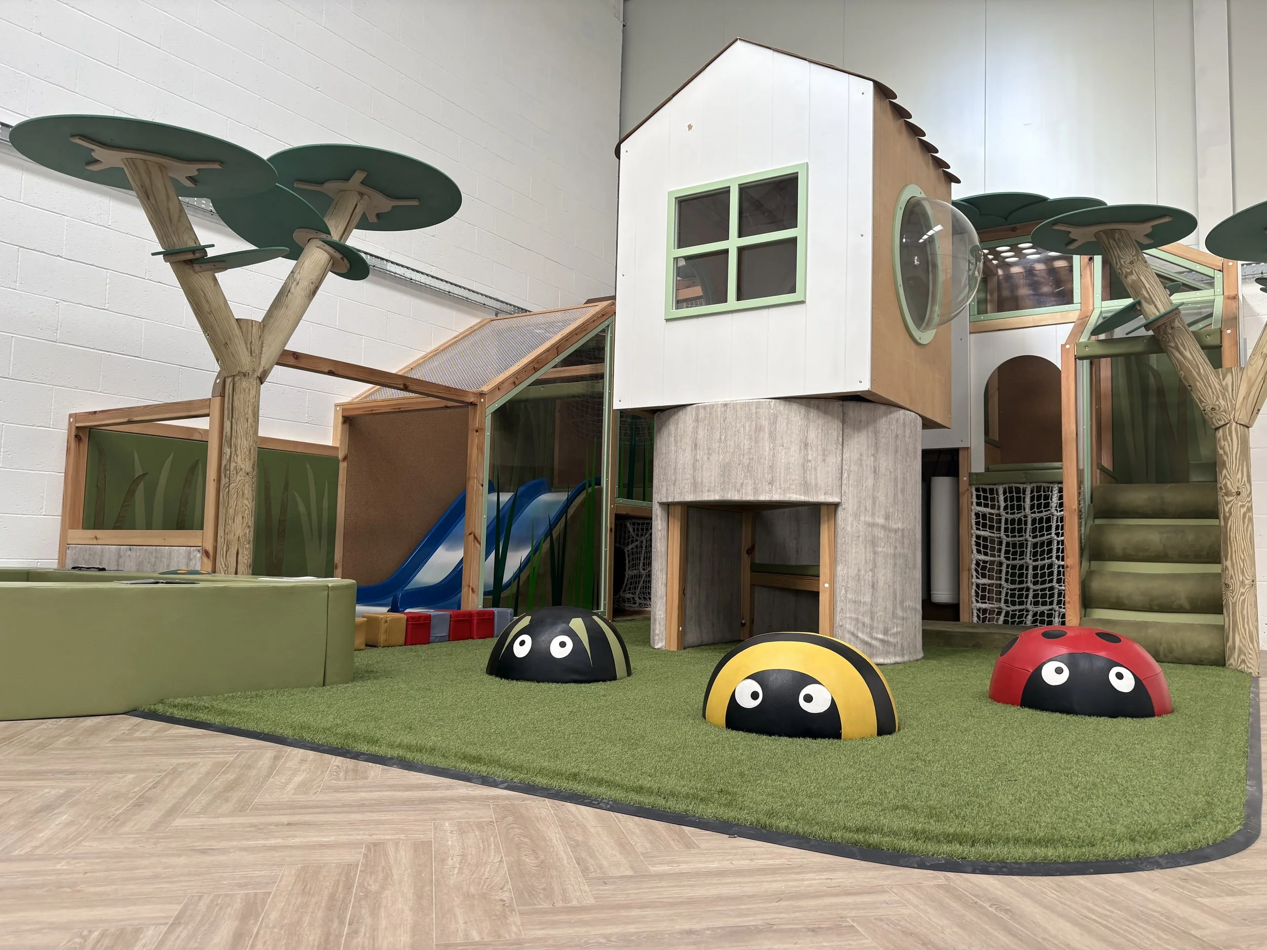 Soft play cafe showing an elevated wooden house surrounded by artifical grass with soft bugs (ladybirds) on top.