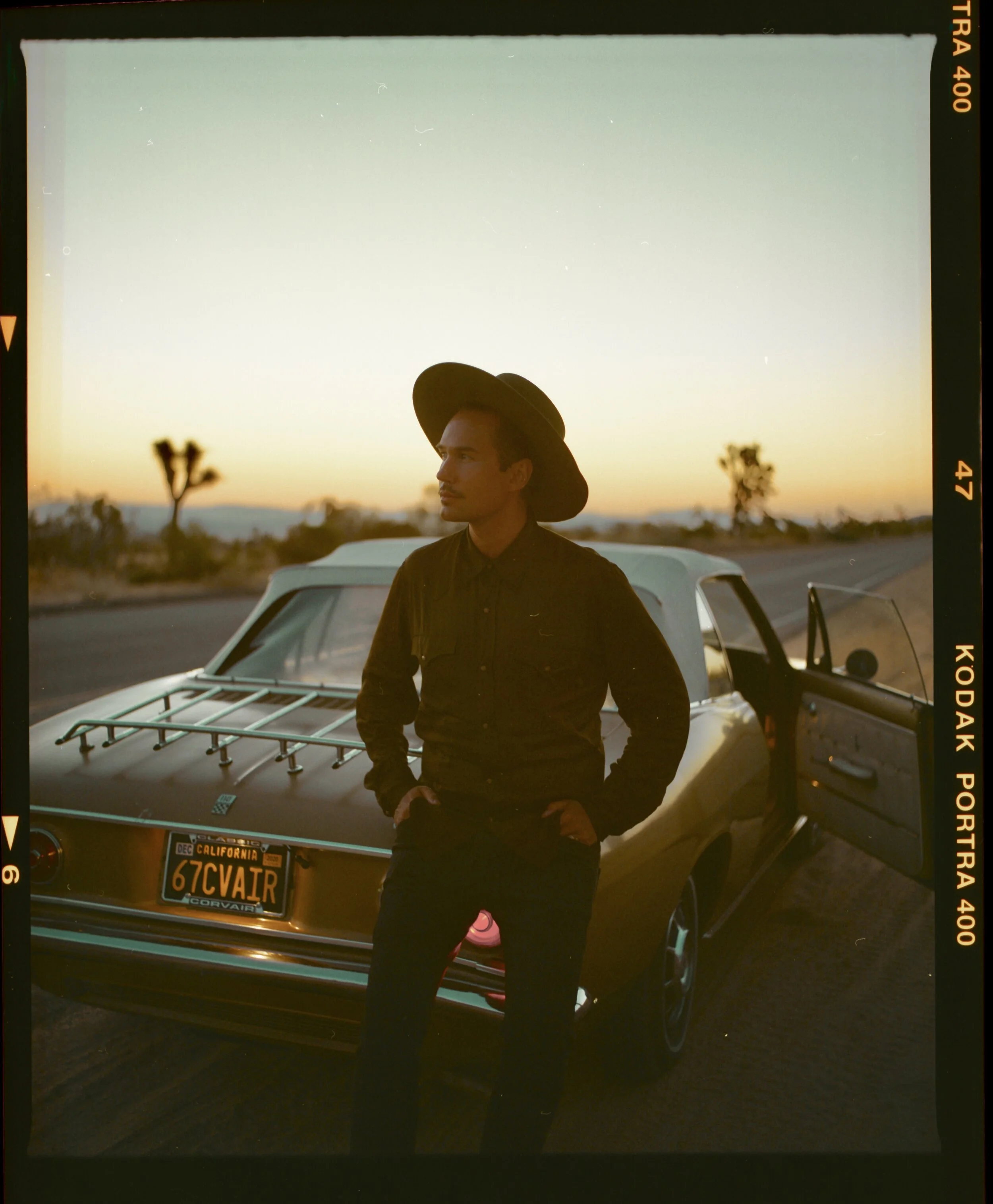 Jonas Myrin wearing a wide-brimmed hat and dark clothing stands next to an open car door on a desert road at sunset, with a vintage station wagon behind him and desert landscape with Joshua trees in the background.