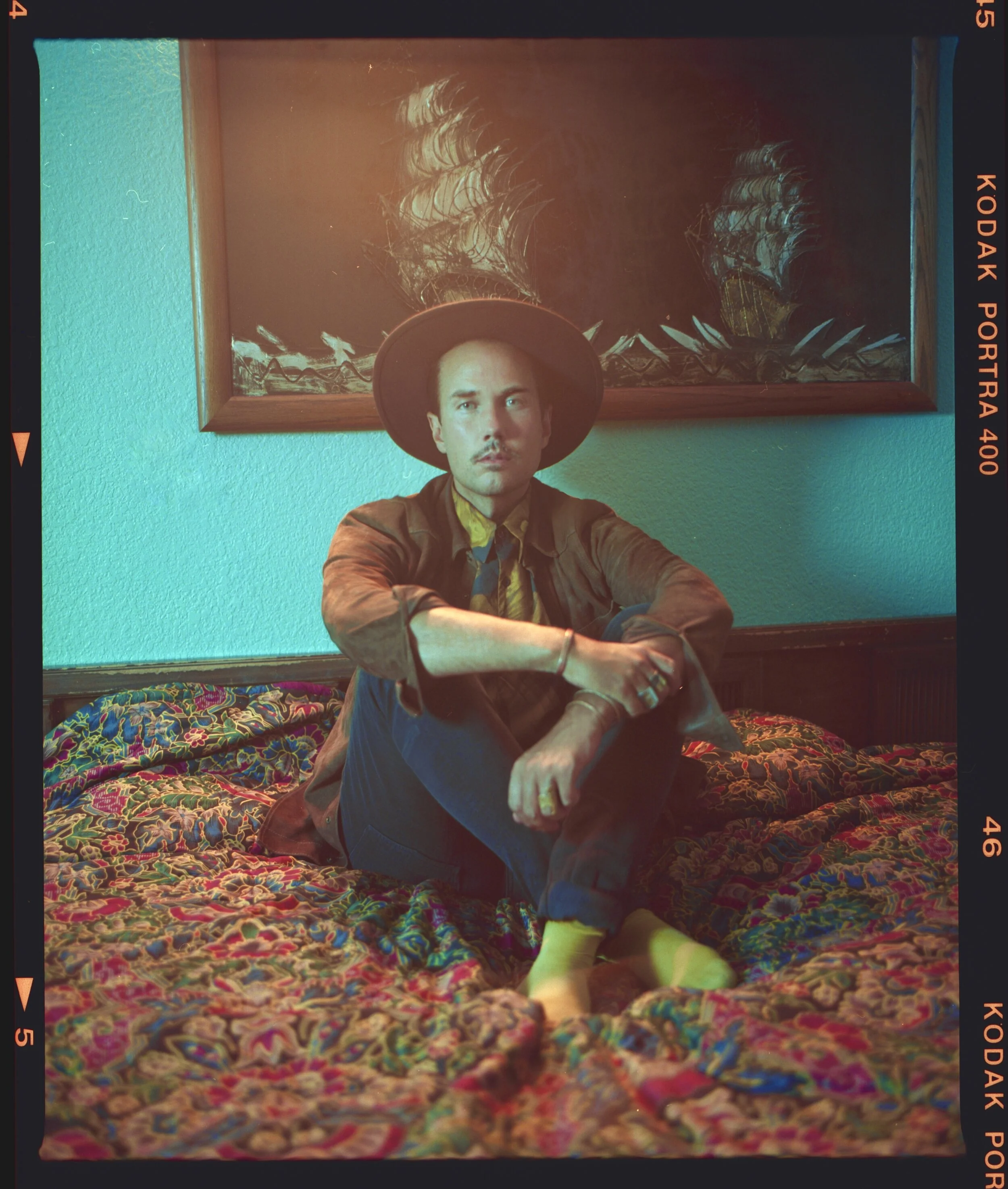 Jonas Myrin sitting on a bed with colorful, intricate bedding, wearing a wide-brimmed hat, a brown jacket, and a yellow shirt, with a serious expression, against a green wall with a framed painting of boats.