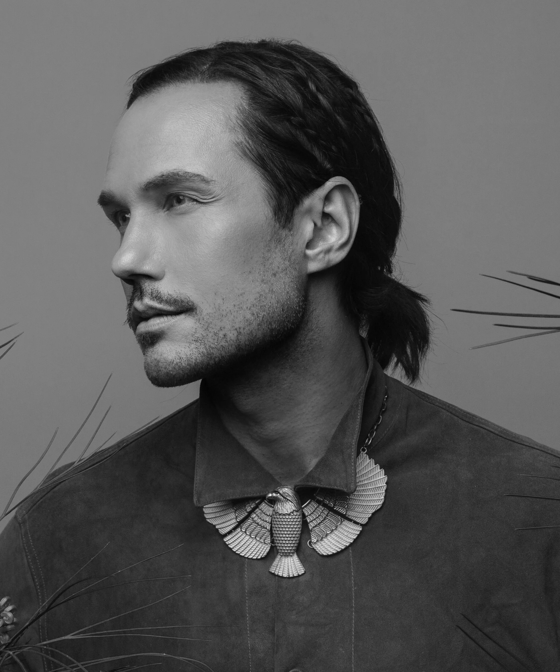 A black-and-white portrait of Jonas Myrin with long hair, dressed in a collared shirt, wearing an ornate, butterfly-shaped necklace.