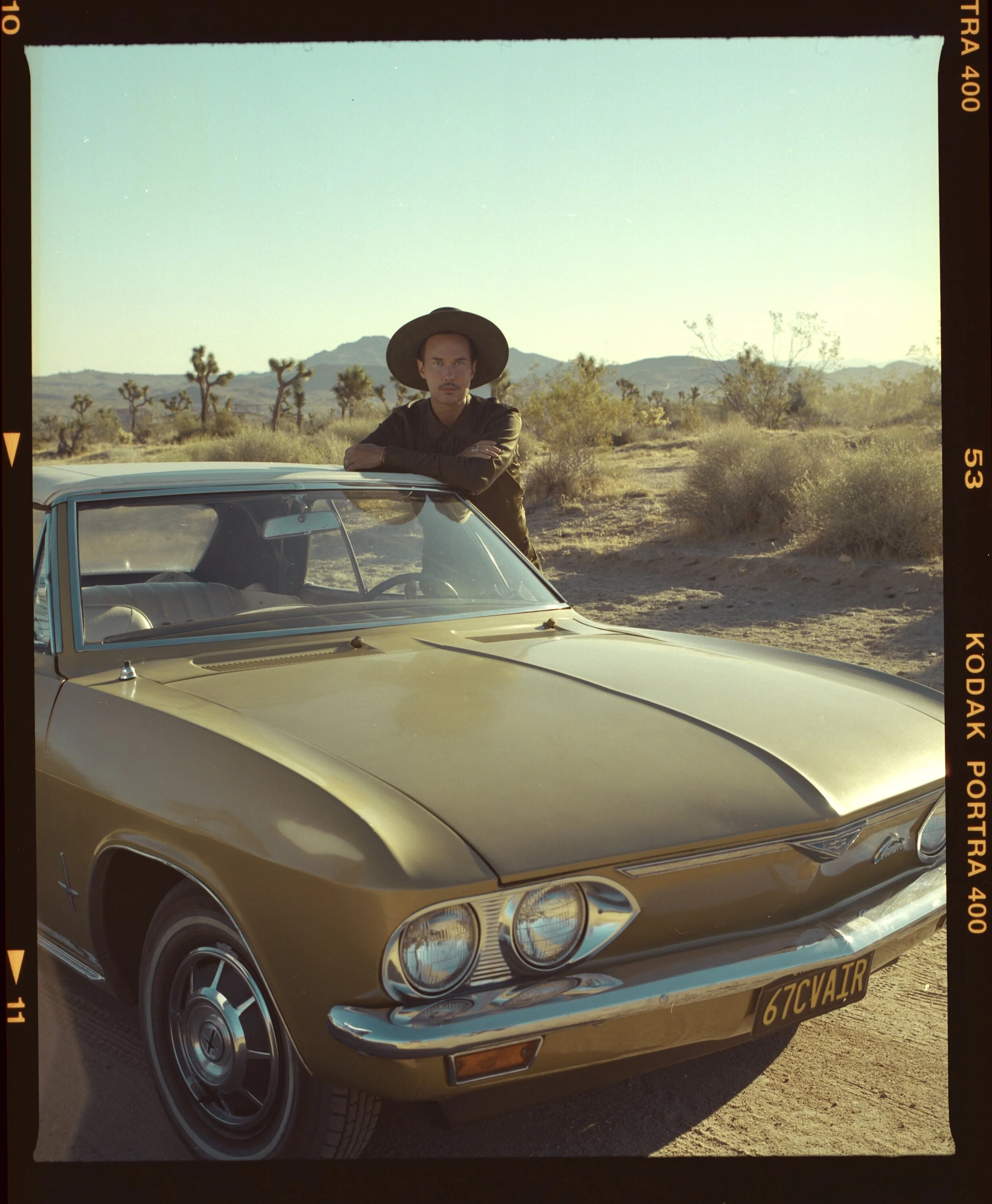 Jonas Myrin wearing a hat leaning on a vintage yellow car in a desert landscape with mountains and Joshua trees in the background.