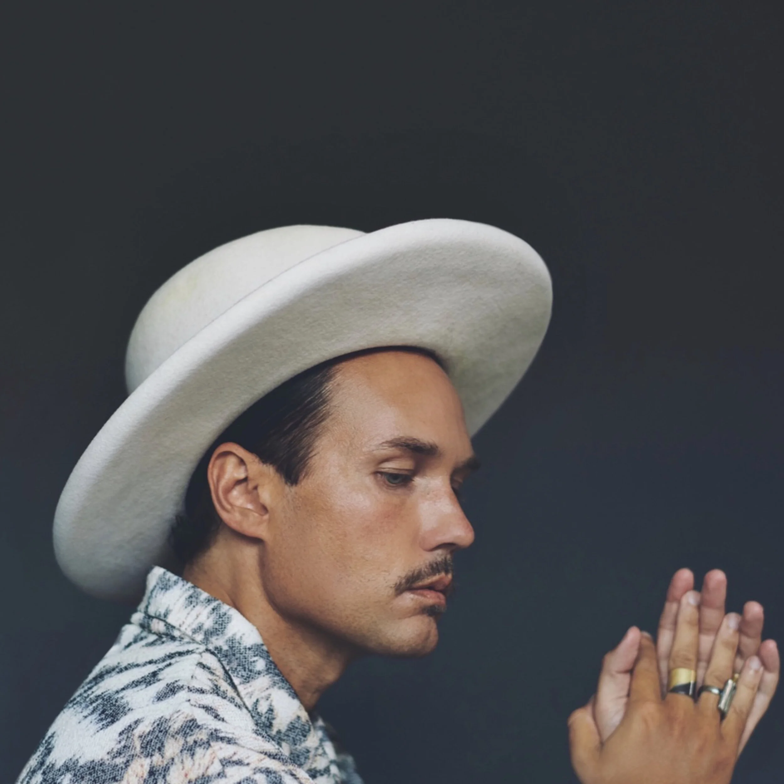 Jonas Myrin, wearing a white cowboy hat and patterned shirt, with a moustache and rings on his fingers, is shown in a side profile, eyes closed, hands together in a prayer-like gesture against a dark background.