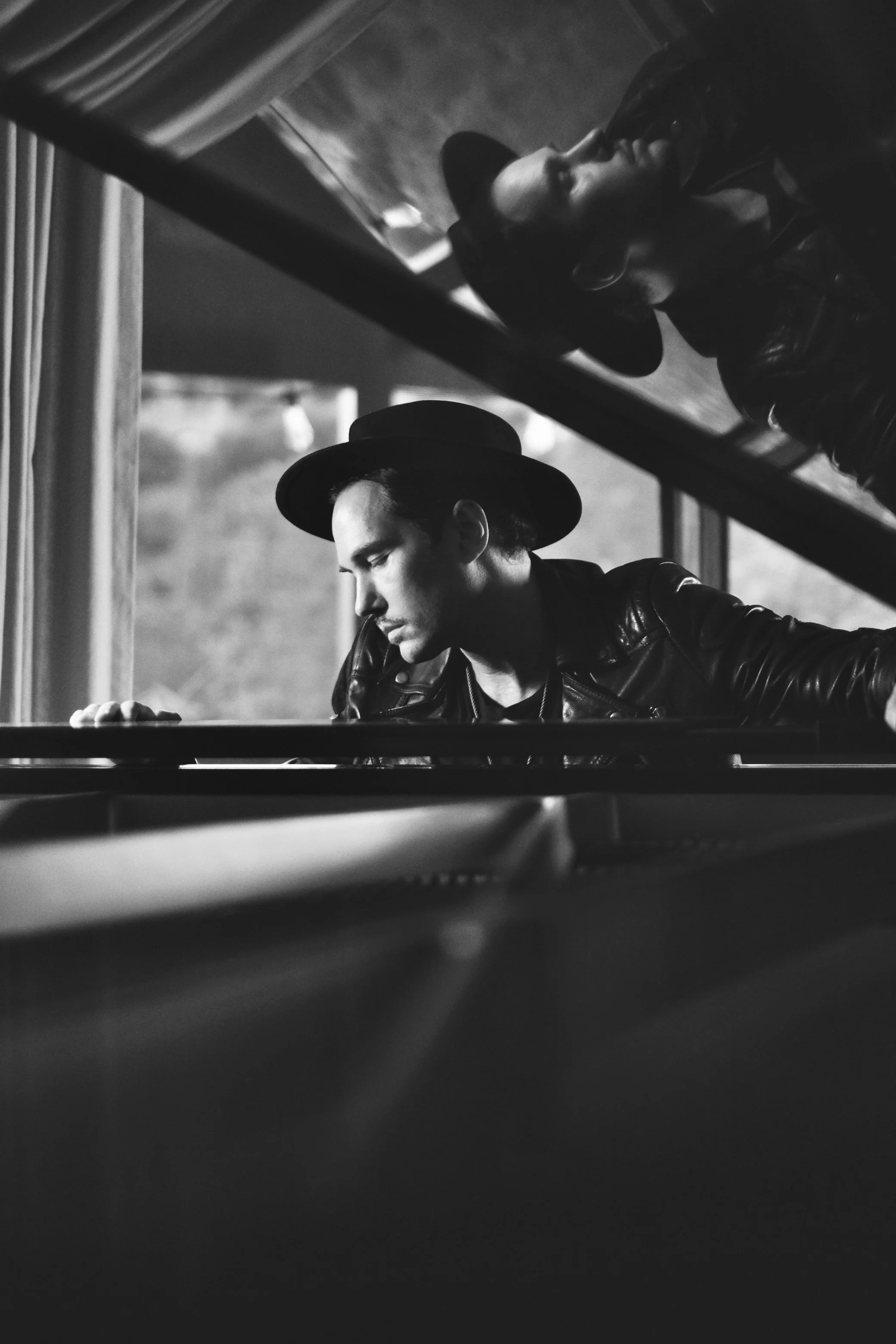 A black and white photo of a young man wearing a hat and leather jacket, playing a grand piano, with his reflection visible on the piano's surface.