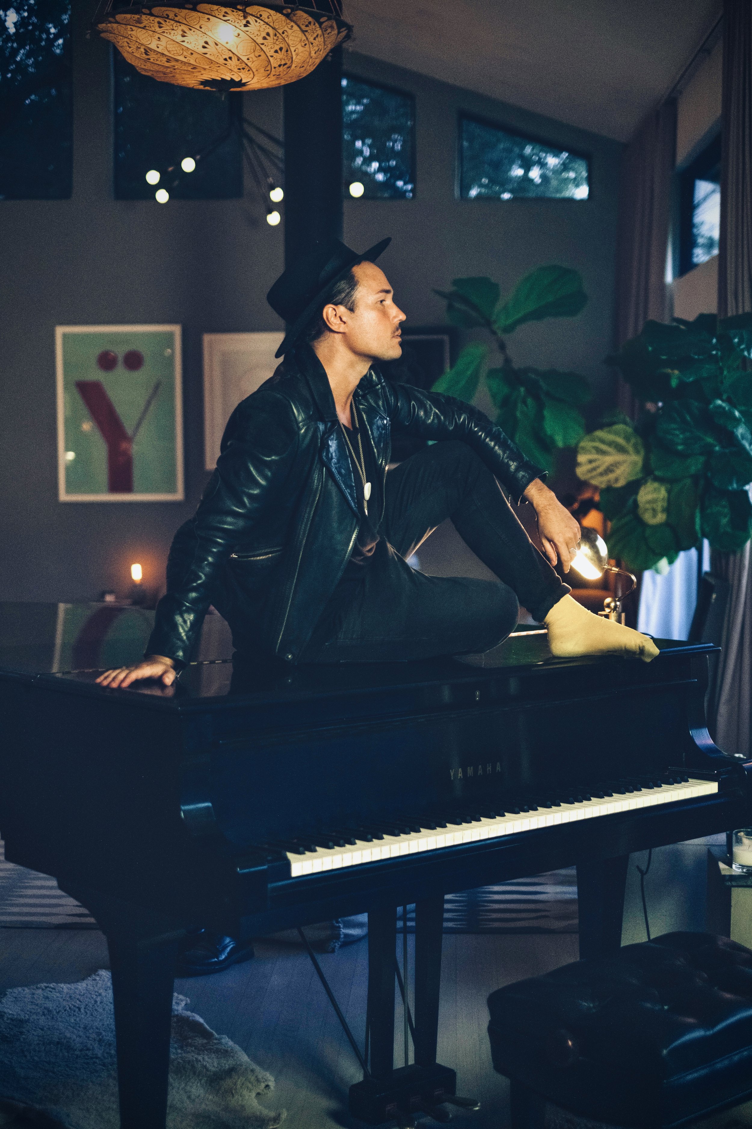 Songwriter Jonas Myrin sitting on top of a grand piano in his Atlas House Studio wearing a hat a leather jacket, black jeans and mustard yellow socks. His shoes can be spotter under the piano.