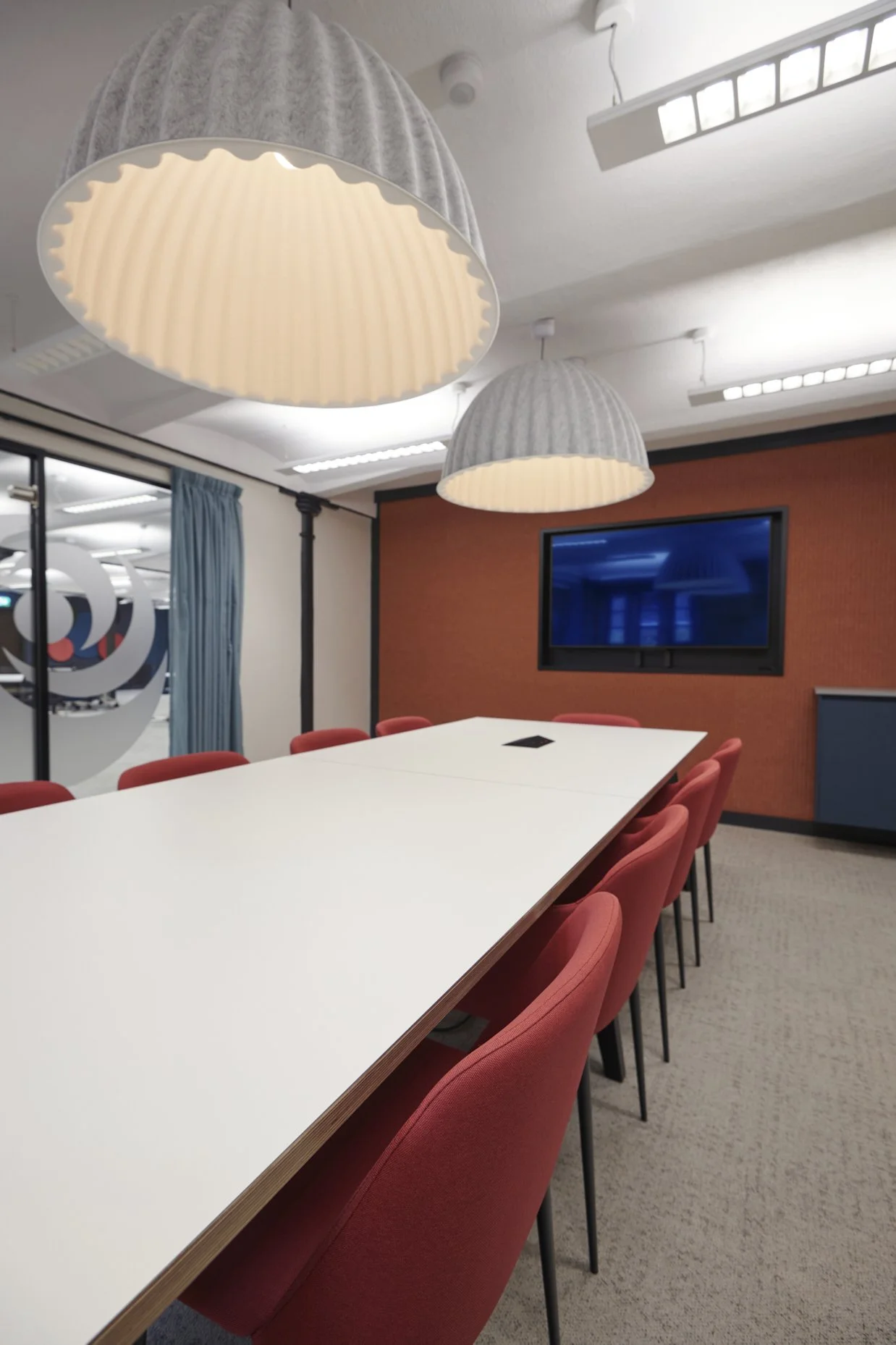 Conference room with a long white table, red chairs, hanging pendant lights, a wall-mounted TV, and a window with a blue curtain.
