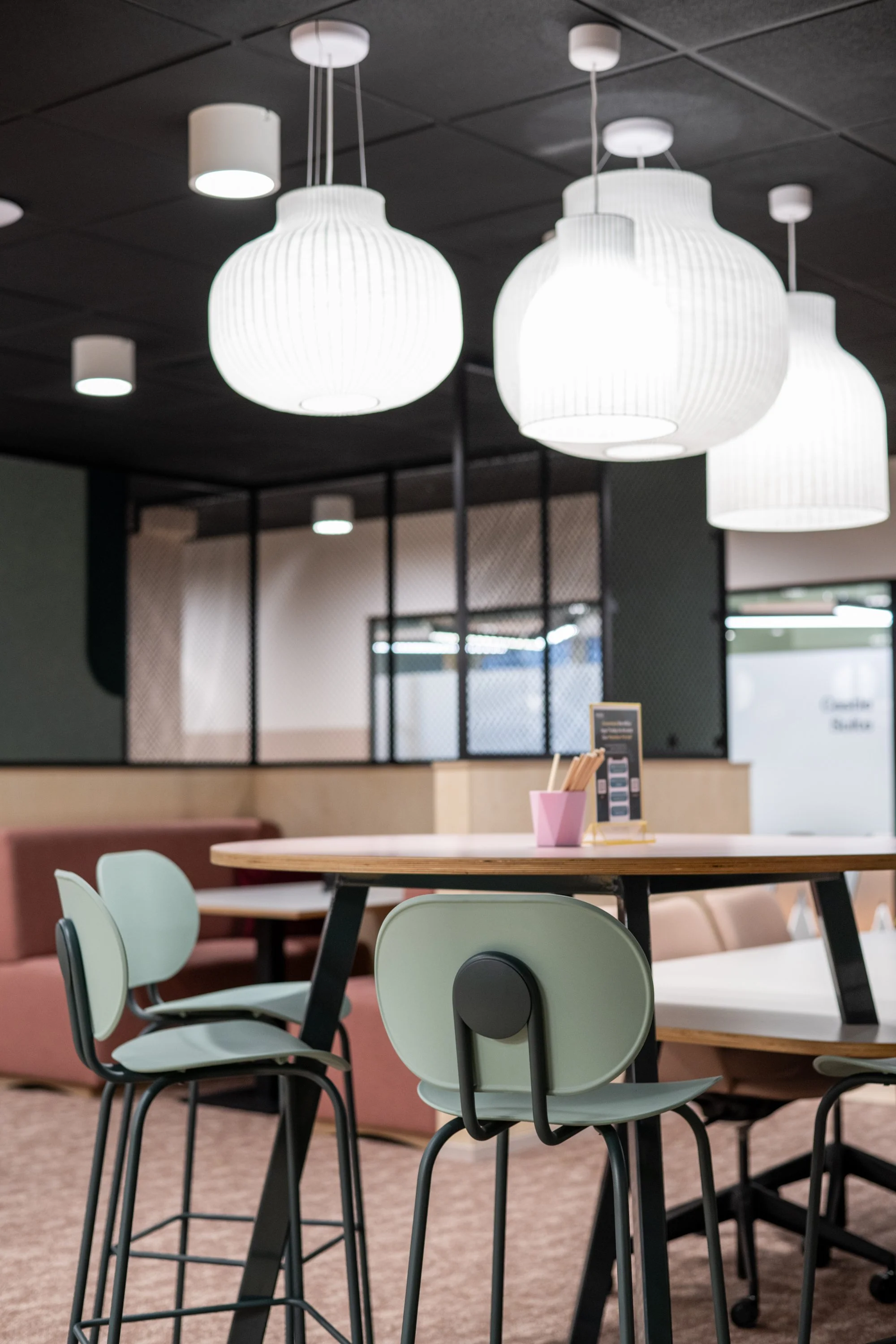 Modern, brightly lit interior with hanging white pendant lights, a wooden table, mint green chairs, and a pink cup with utensils on the table
