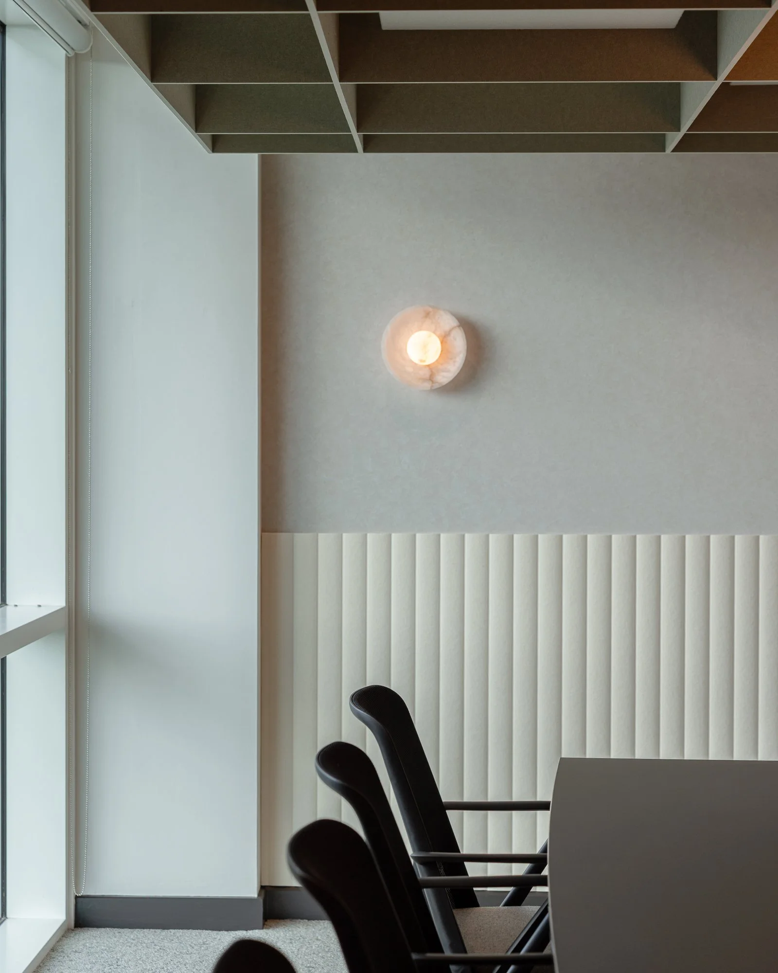 Modern office boardroom space with a row of black chairs, a black desk, white acoustic half height panelling and a decorative wall light on the wall made from Alabaster