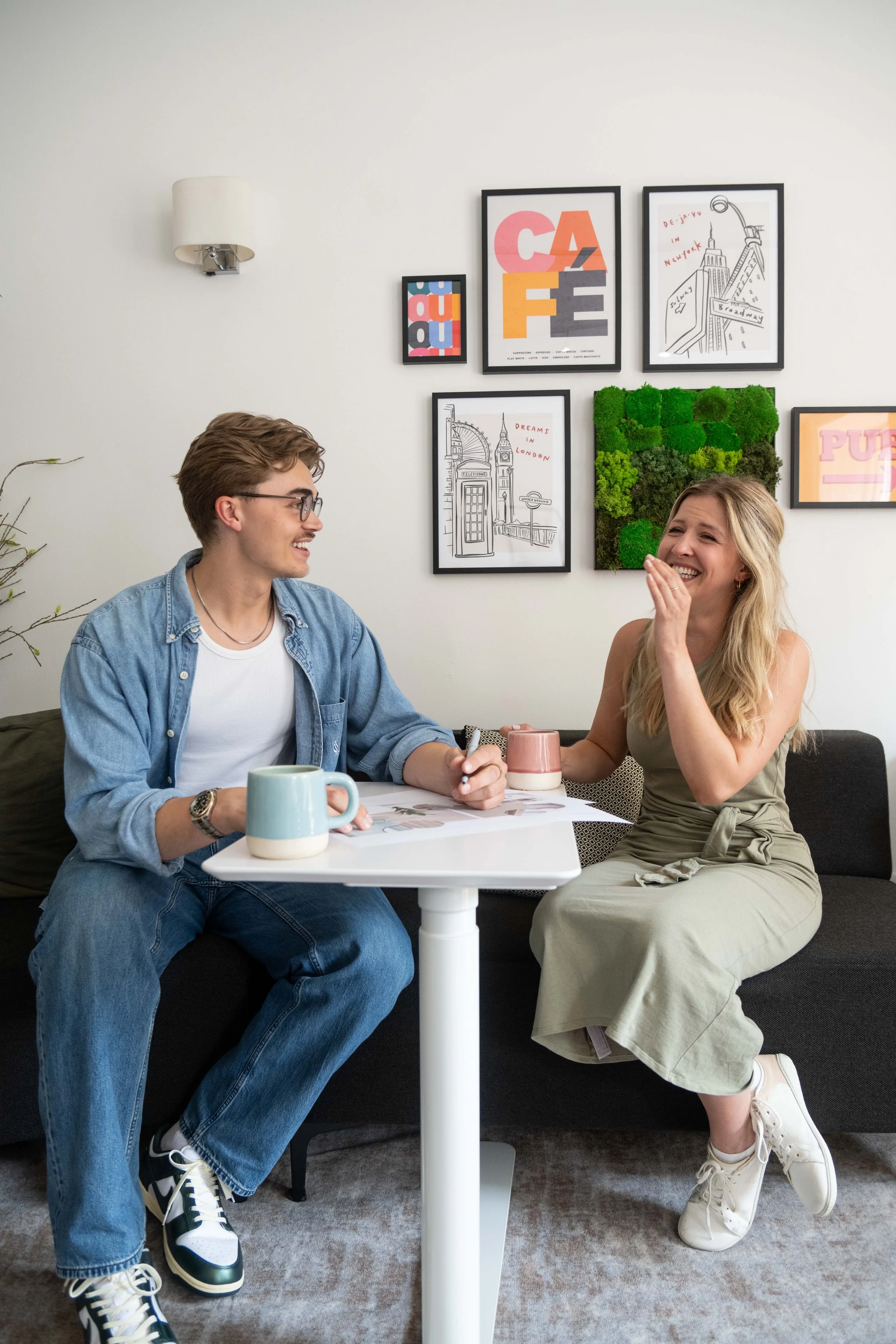 A young man and woman sitting at a small white table, laughing and talking in a living room with framed art on the wall, mugs on the table, and a sofa.
