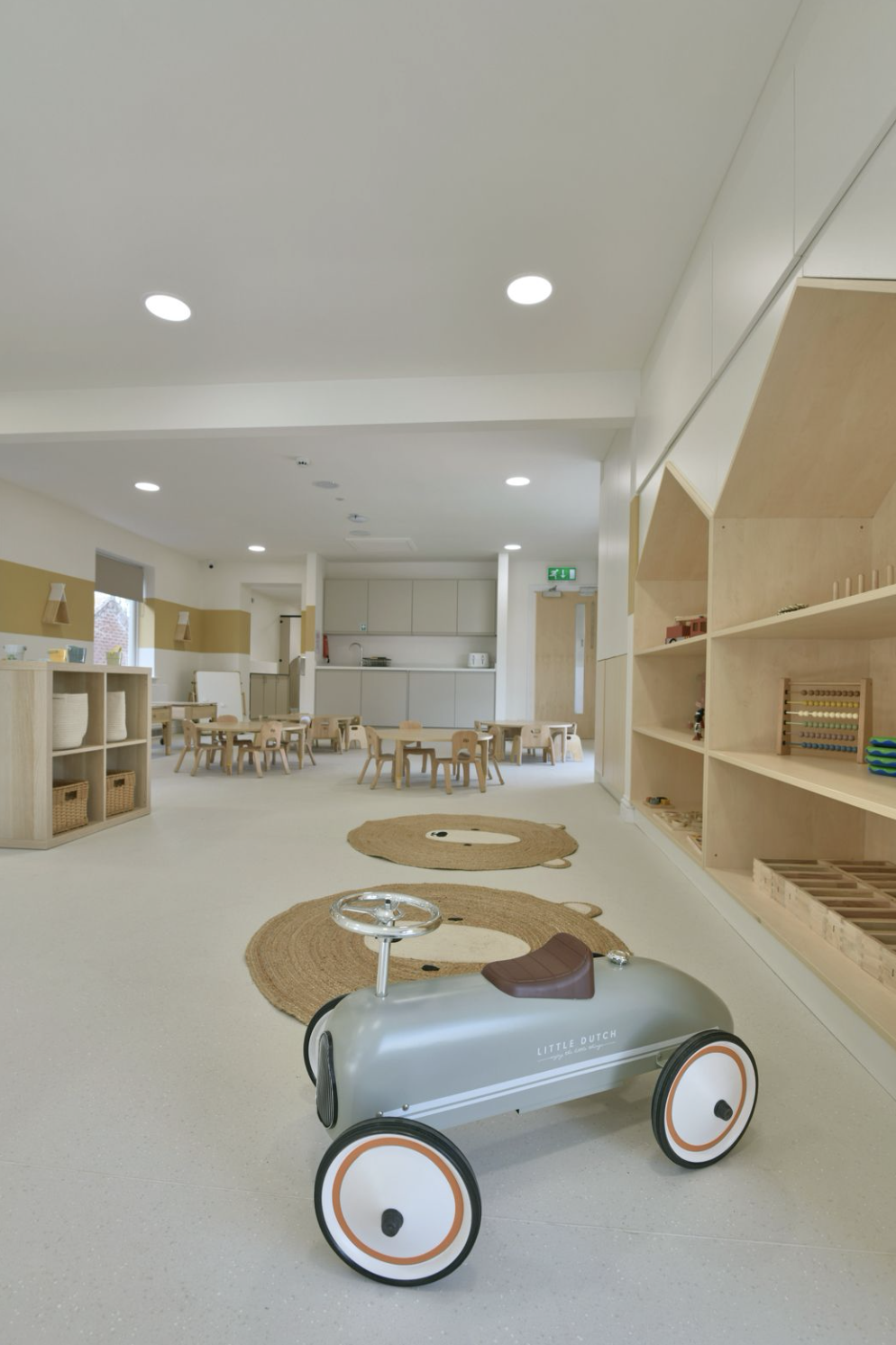 A spacious, modern preschool classroom with natural wood furniture, small tables and chairs, toys, and educational materials. There are two rugs and a ride-on toy in the foreground, with soft lighting and an overall neutral color palette.