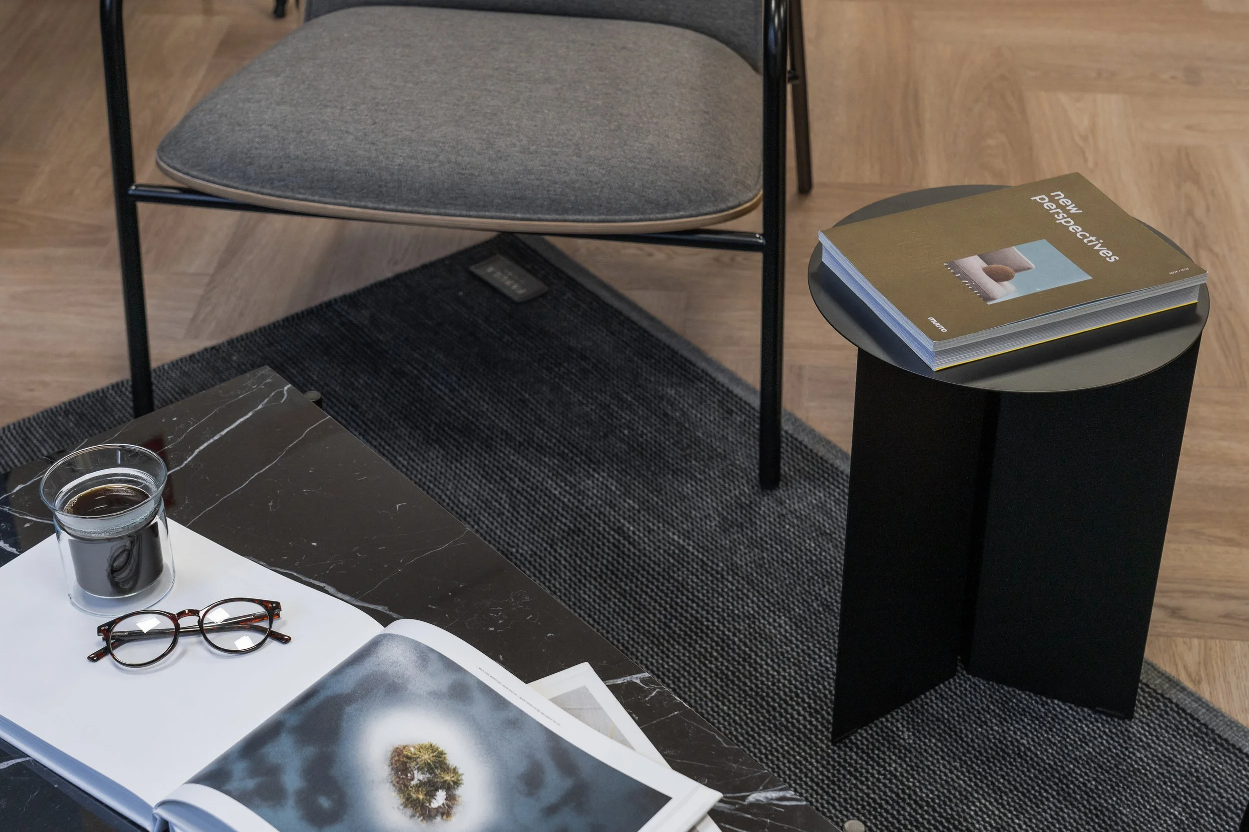 A black and white marble coffee table with an open magazine, a pair of round glasses, and a small glass cup of coffee. In the background is a gray chair with a black metal frame, a black pedestal with a magazine titled 'new perspectives,' and a black rug on a wooden floor.